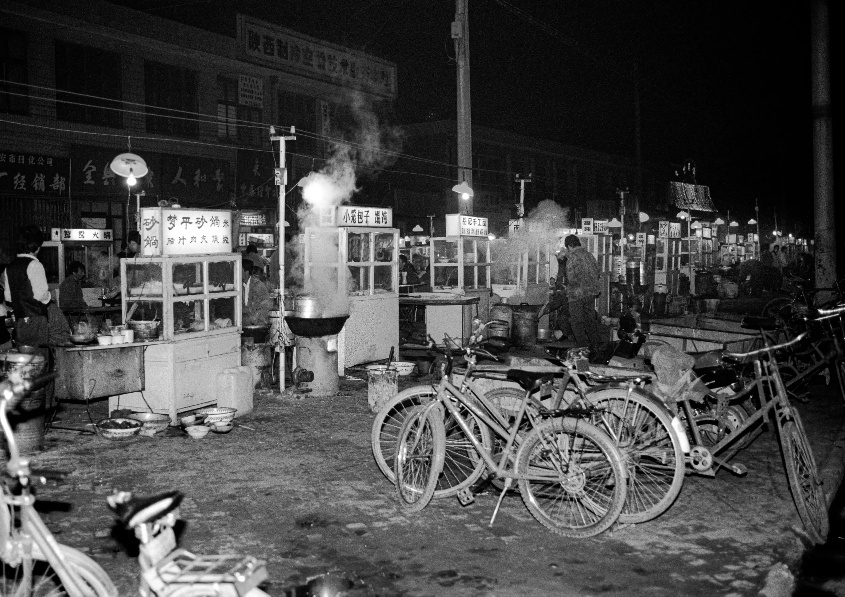 Une rue de nuit en Chine avec des vélos au premier plan, des échoppes fumantes au centre et une succession de stands éclairés vers le fond