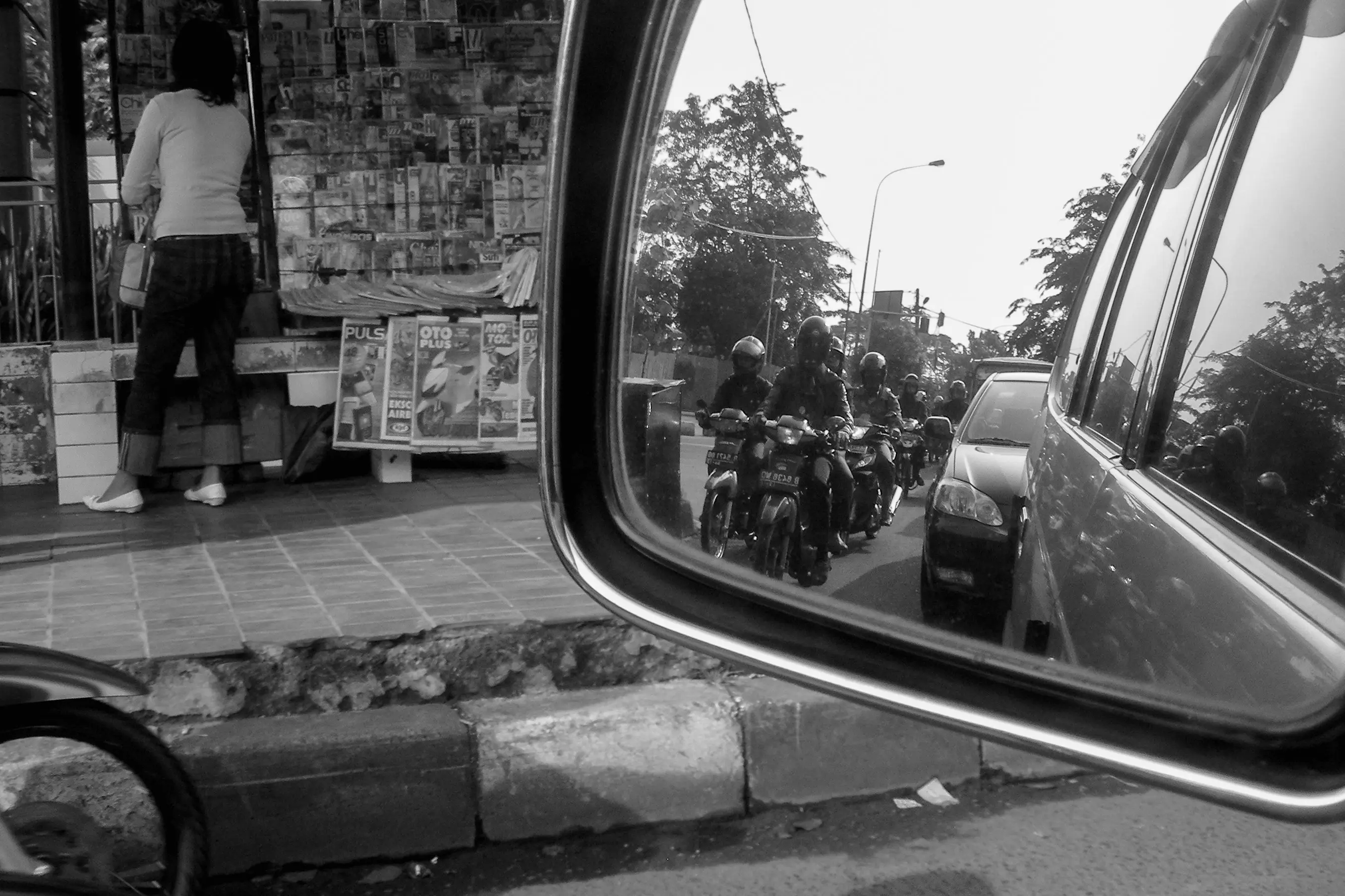 Dans un rétroviseur apparaissent des motos et une voiture, tandis que le bord de la route et un kiosque occupent le champ principal