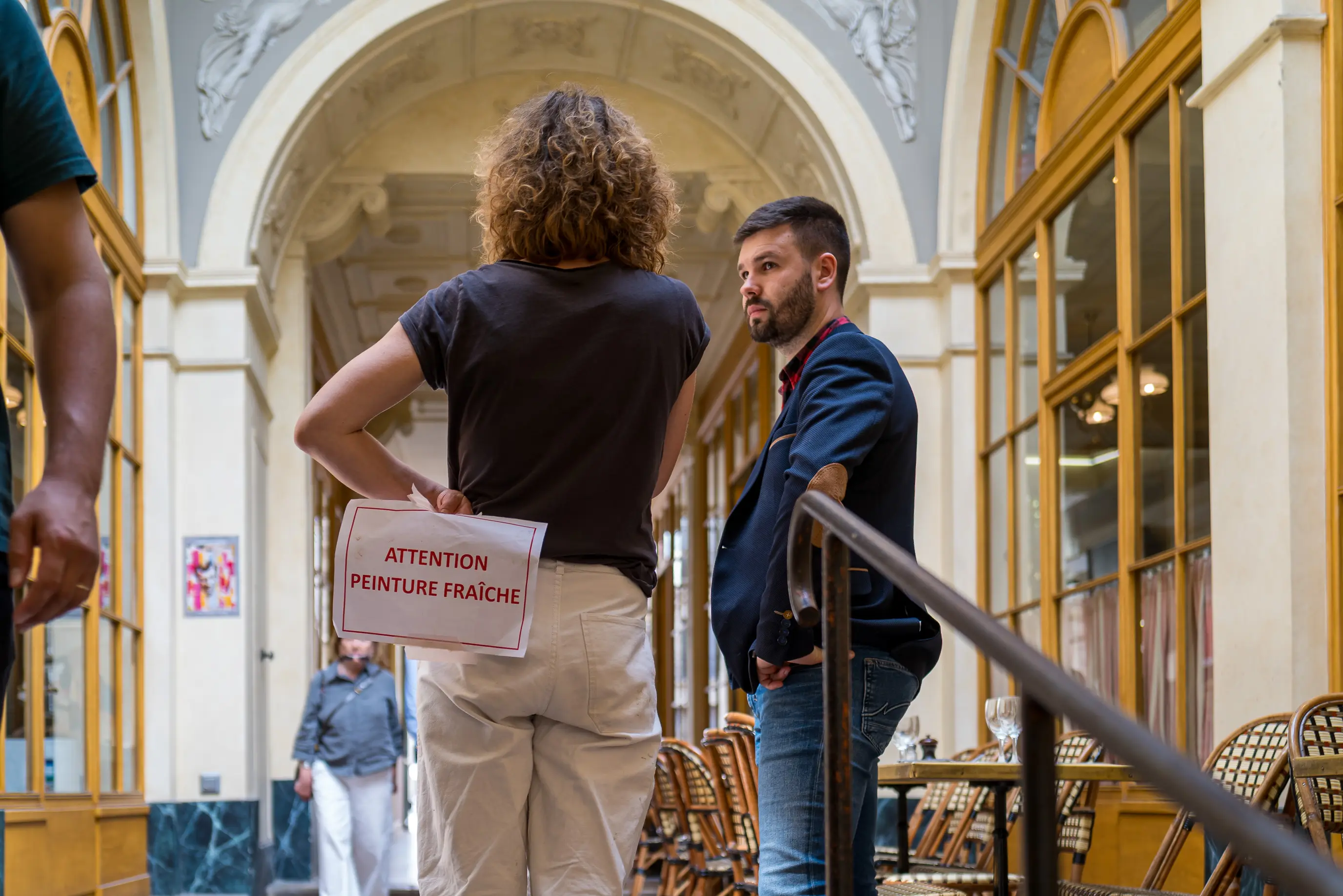 Une femme tient derrière elle une pancarte portant l'inscription Attention peinture fraîche tandis qu'elle parle avec un homme