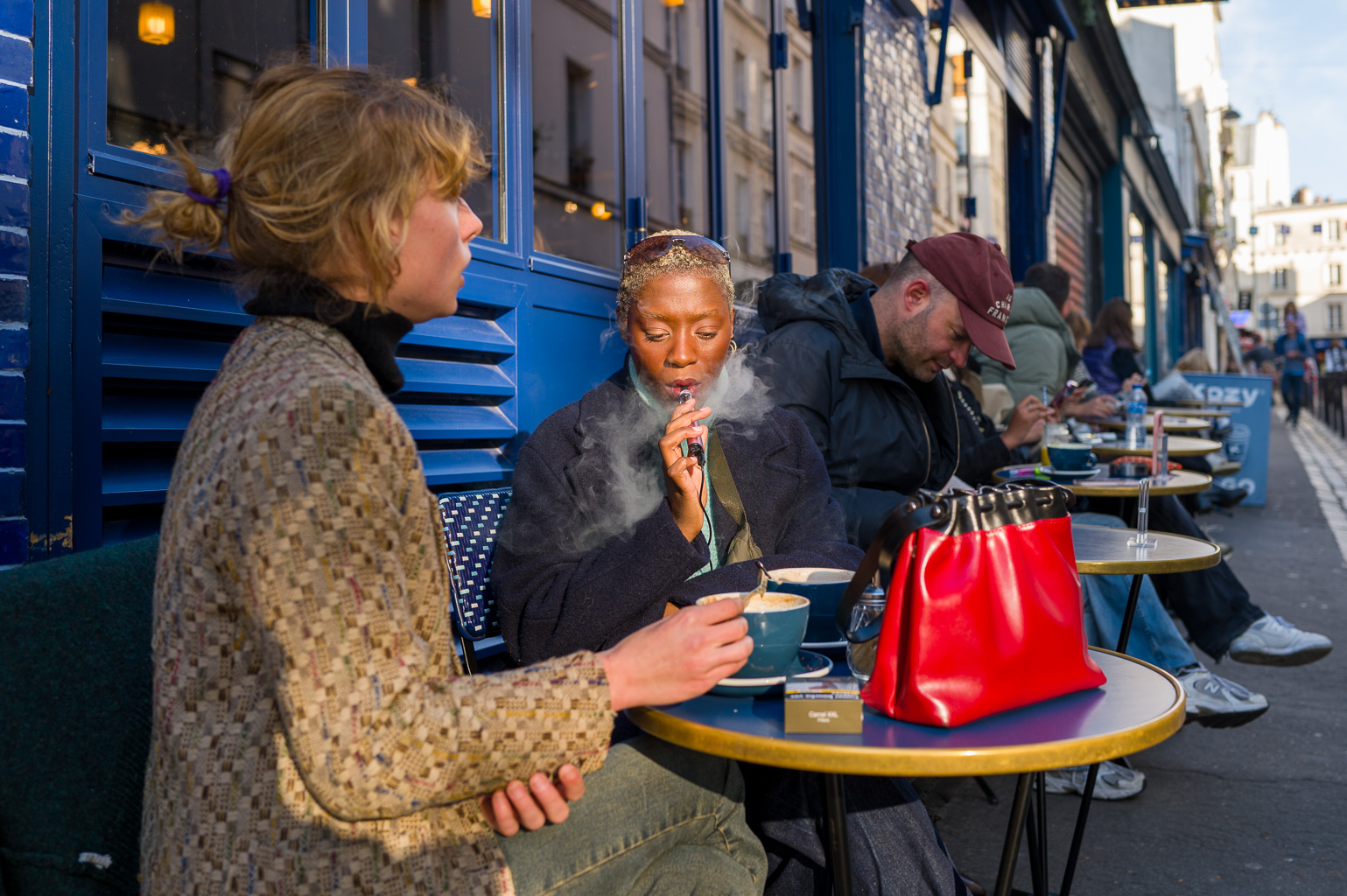 Deux femmes discutant à la terrasse d’un café parisien, l’une vapotant dans la lumière du matin, l’autre tenant sa tasse, sur fond de façade bleue et d’ambiance détendue.