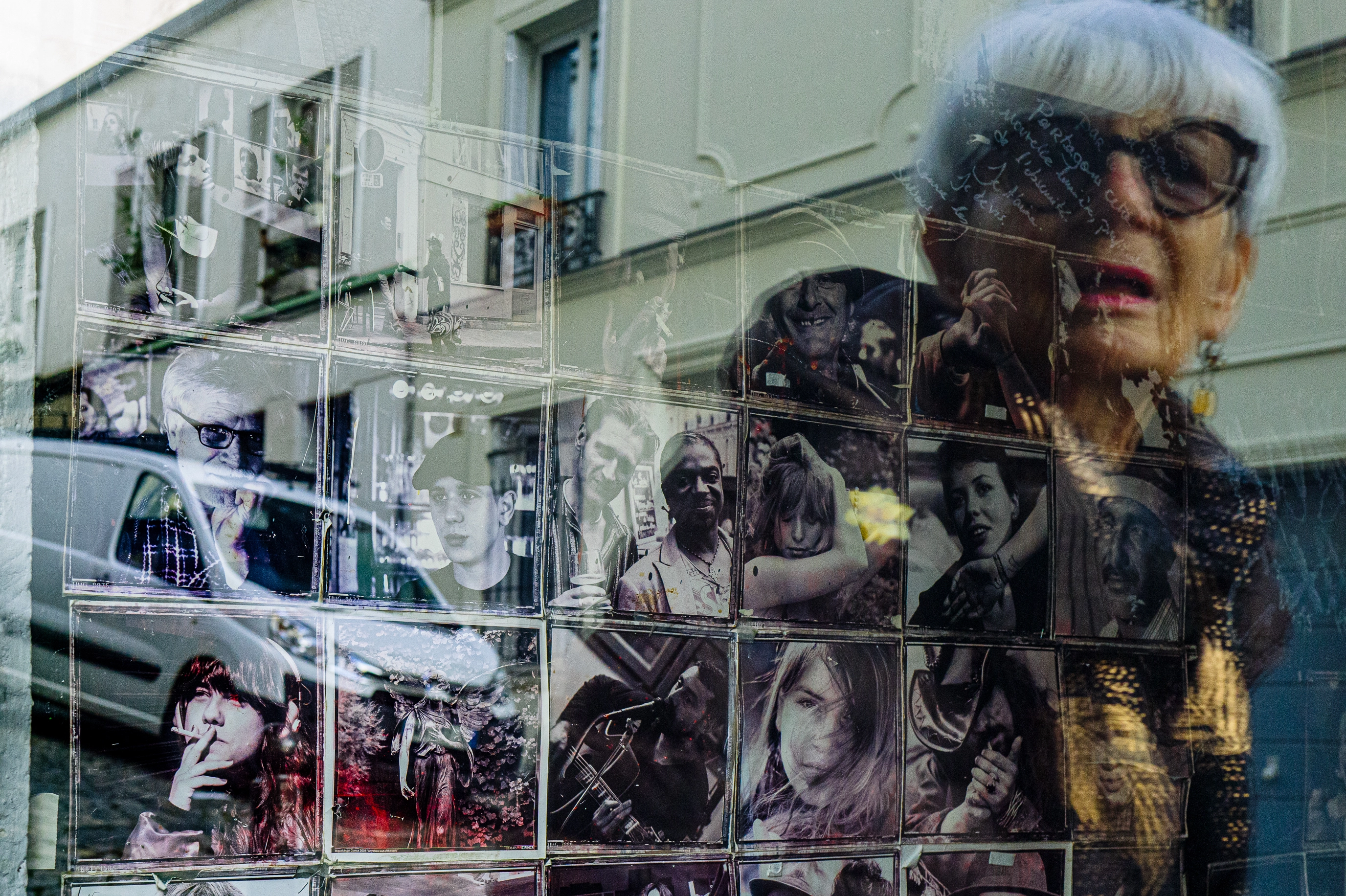Des portraits sur des carreaux de faïence apparaissent derrière une vitrine où se reflètent des immeubles et le visage d’une femme qui les regarde