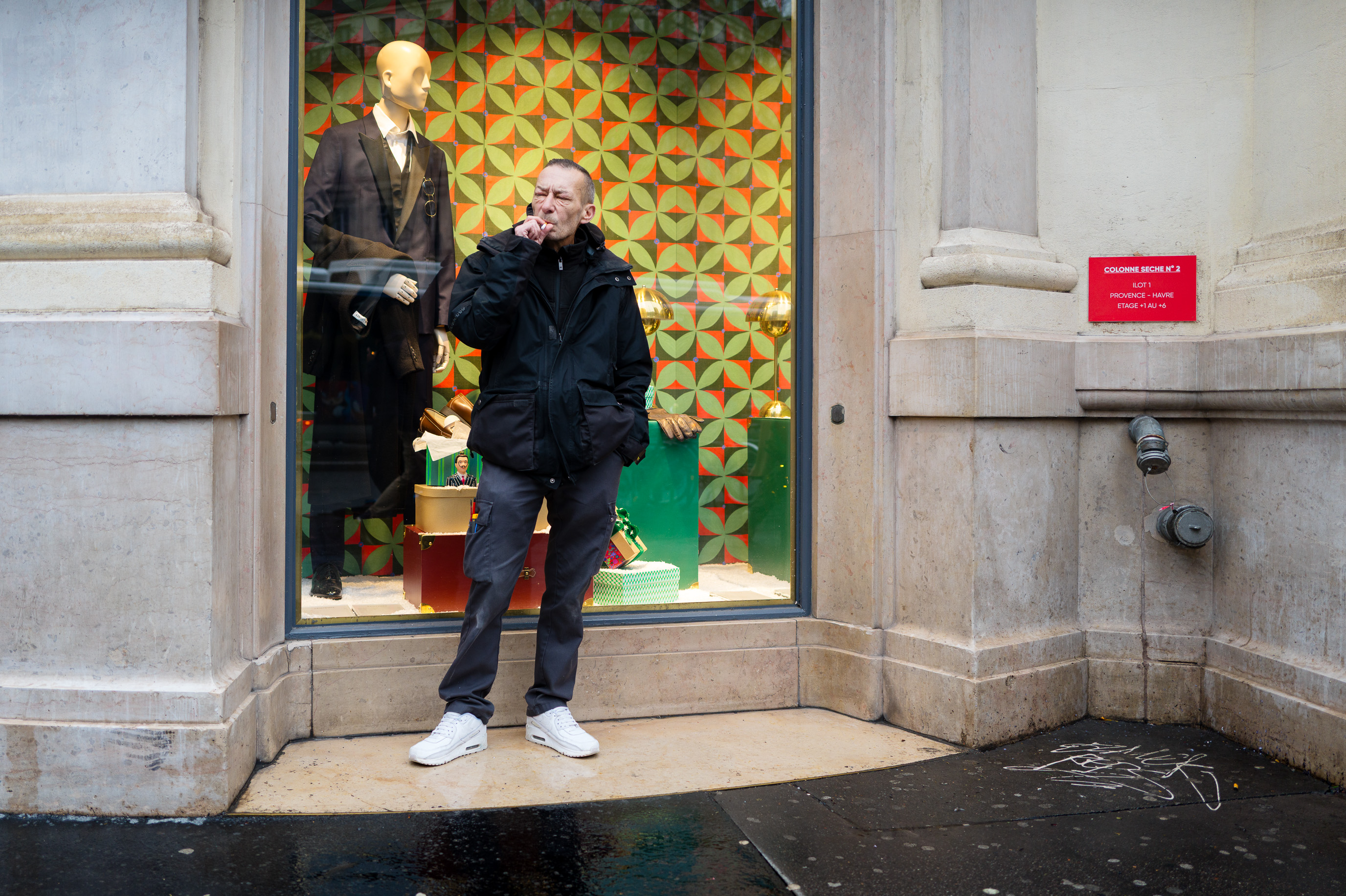 Un homme fume devant la vitrine d'une boutique où un mannequin en costume semble l'observer. Une photographie de rue prise à Paris.