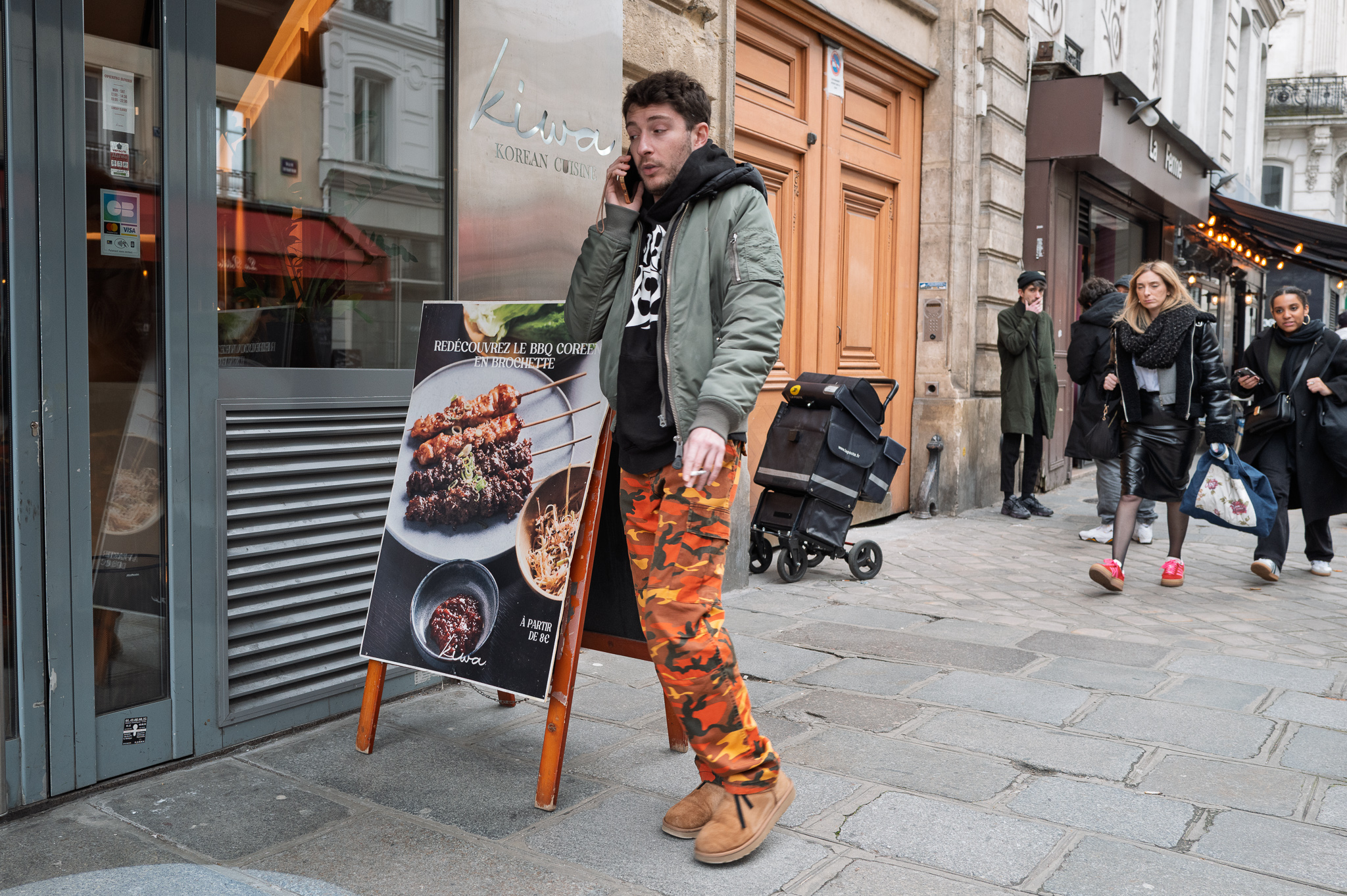 Un homme en pantalon camouflage orange téléphone devant un restaurant coréen, une cigarette à la main, l'air indécis.