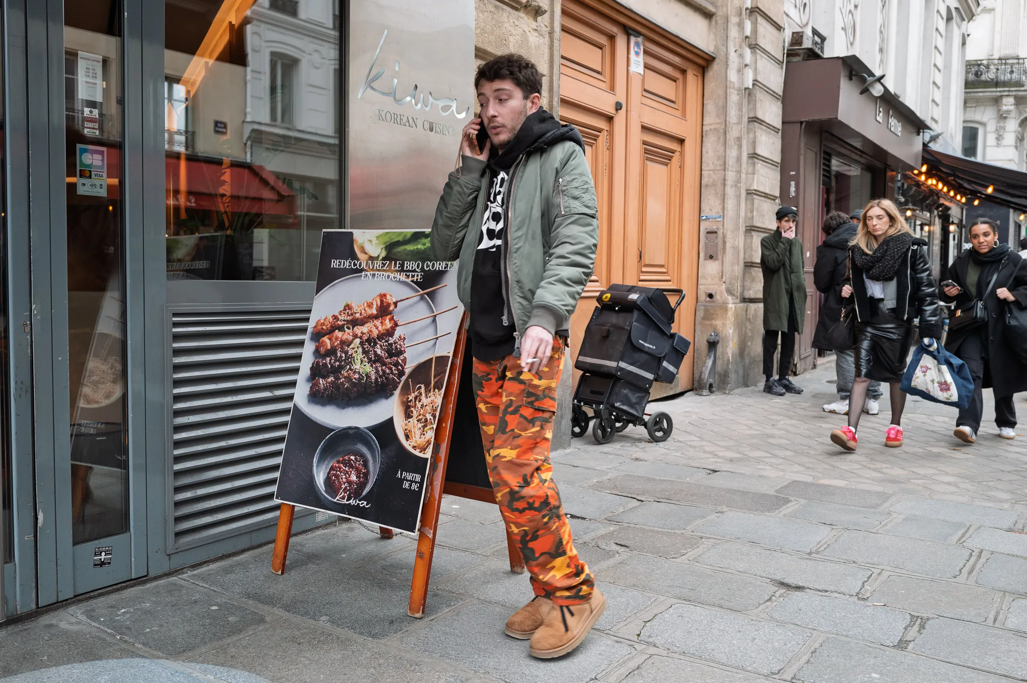 Un homme en pantalon camouflage orange téléphone devant un restaurant coréen, une cigarette à la main, l'air indécis.