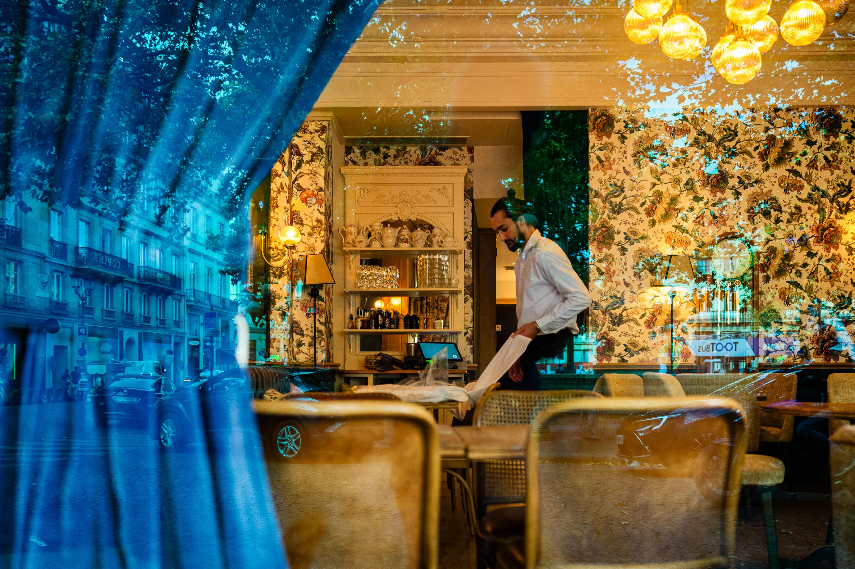 À travers la vitre d'un restaurant, un serveur prépare la salle avant le service tandis que les reflets des arbres et de la rue se mêlent au décor intérieur.