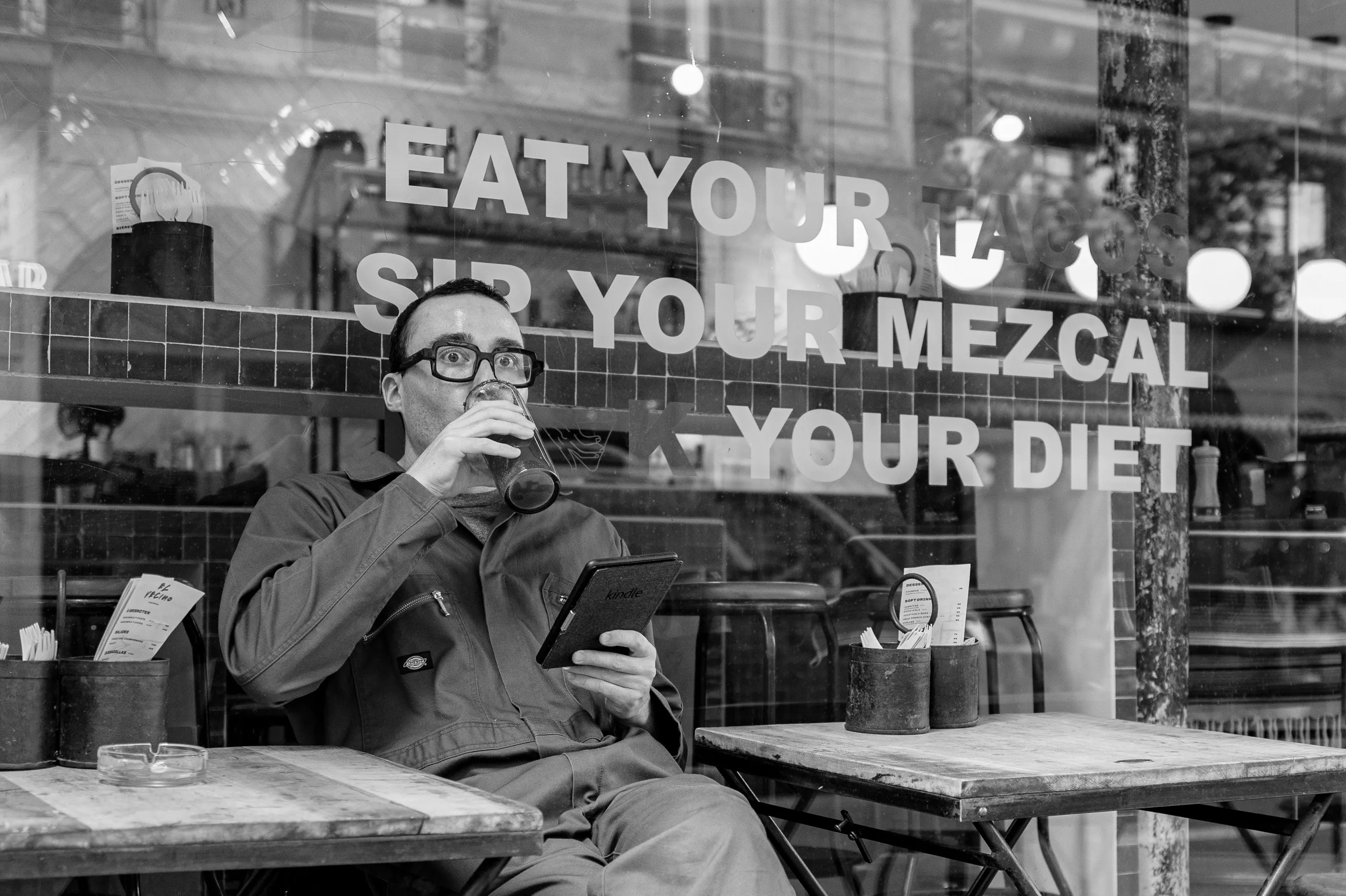 À la terrasse d'un café, un homme interrompt sa lecture sur une Kindle et fixe soudain quelque chose hors champ, un verre suspendu à la main.