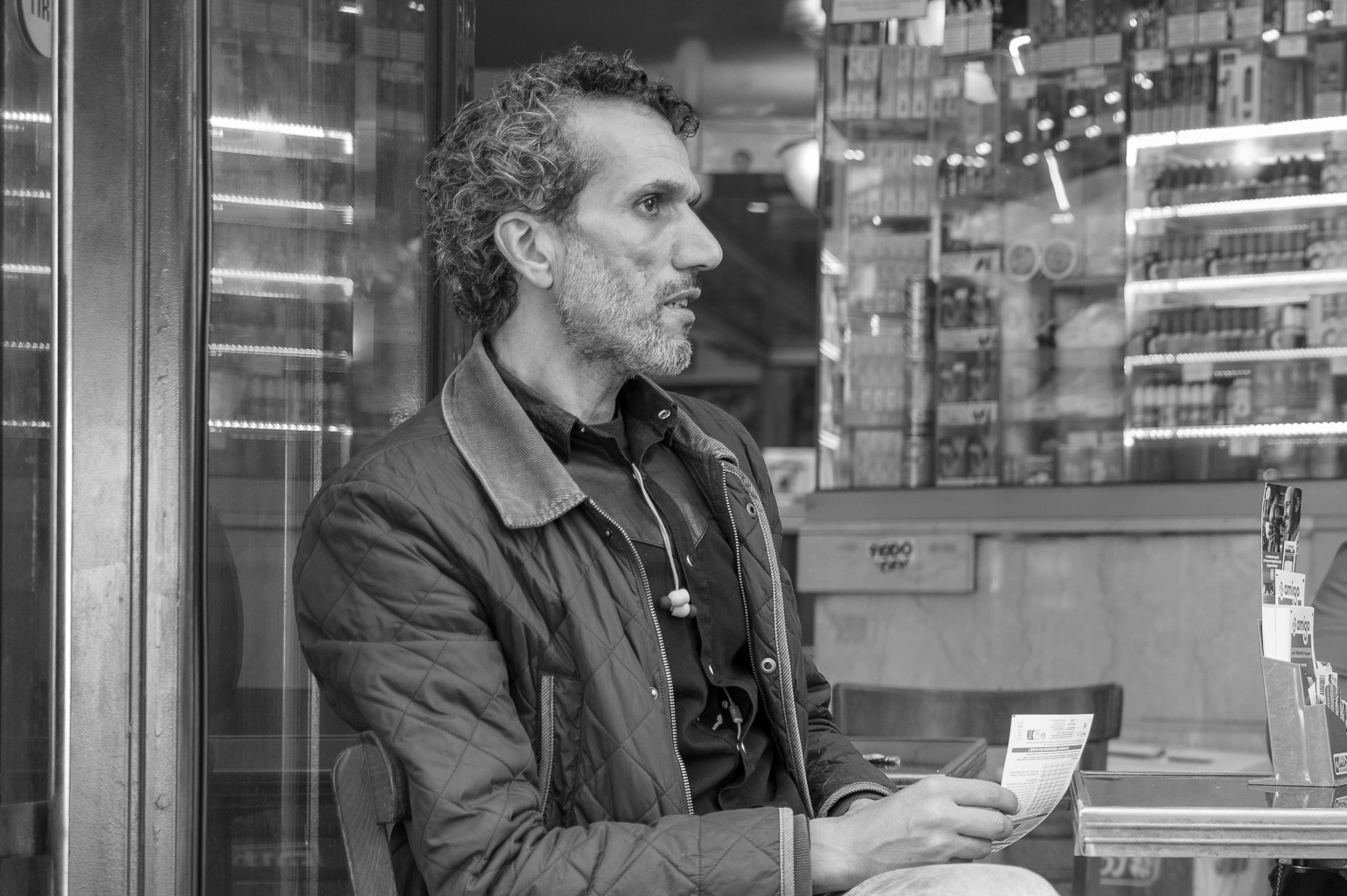 Un homme assis dans un café, tenant un ticket de Loto, entouré d'étagères remplies de bouteilles. Ambiance nostalgique en noir et blanc.