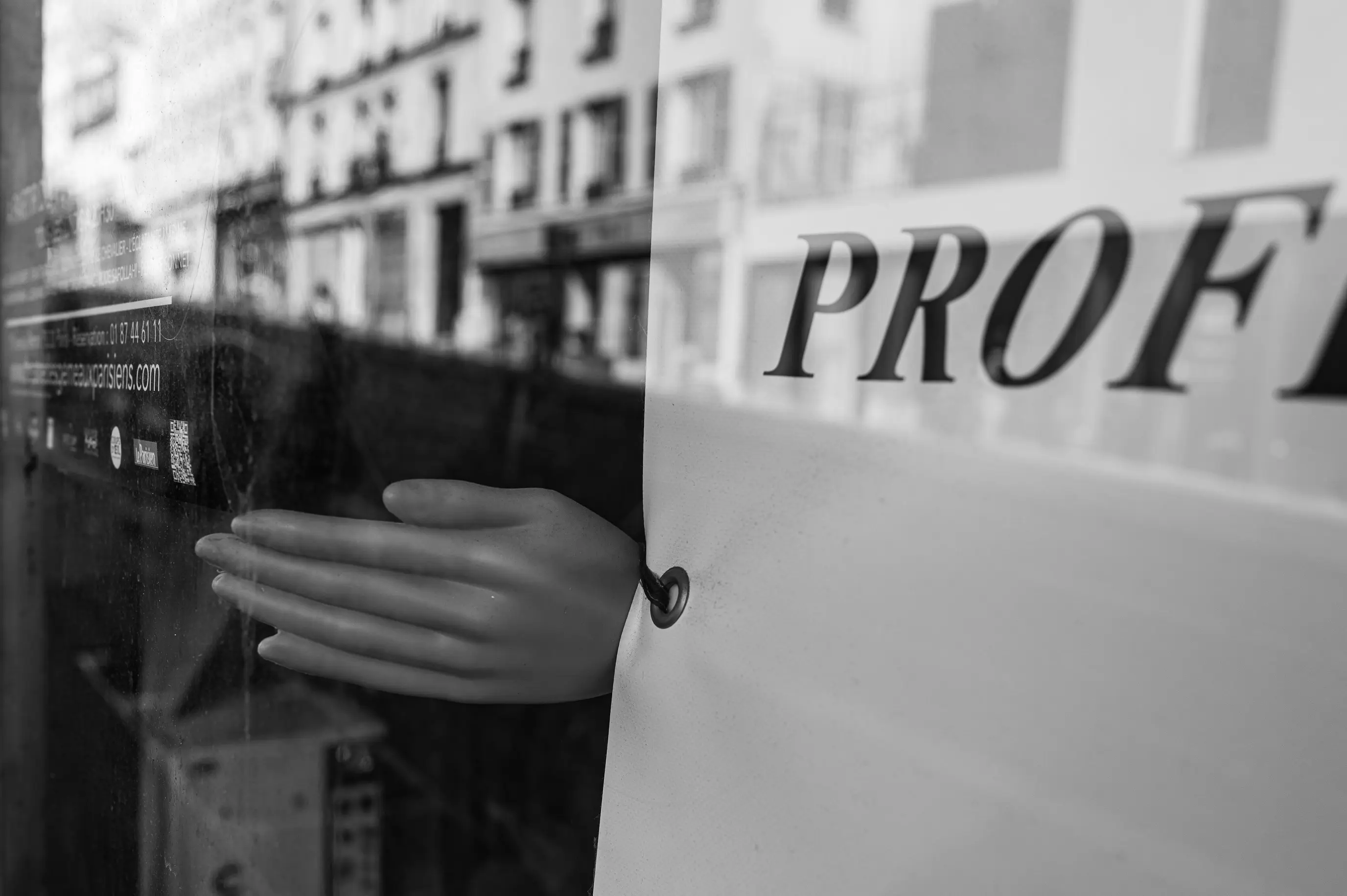 Une main apparaît derrière une vitrine partiellement masquée, parmi les reflets de la rue et une inscription tronquée