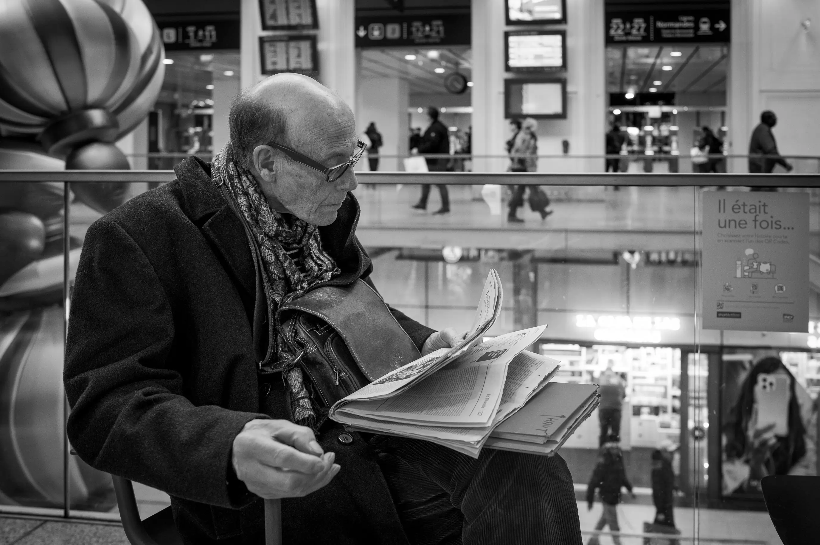 Un homme âgé lit un journal papier dans un lieu de passage, tandis que des passants et des écrans occupent l'arrière-plan