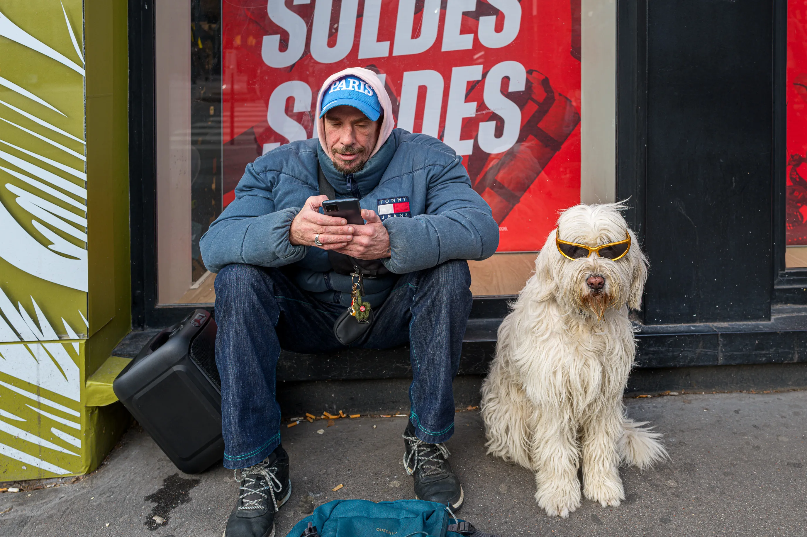 Un homme absorbé par son téléphone est assis à côté de son chien portant des lunettes de soleil devant une vitrine de magasin.