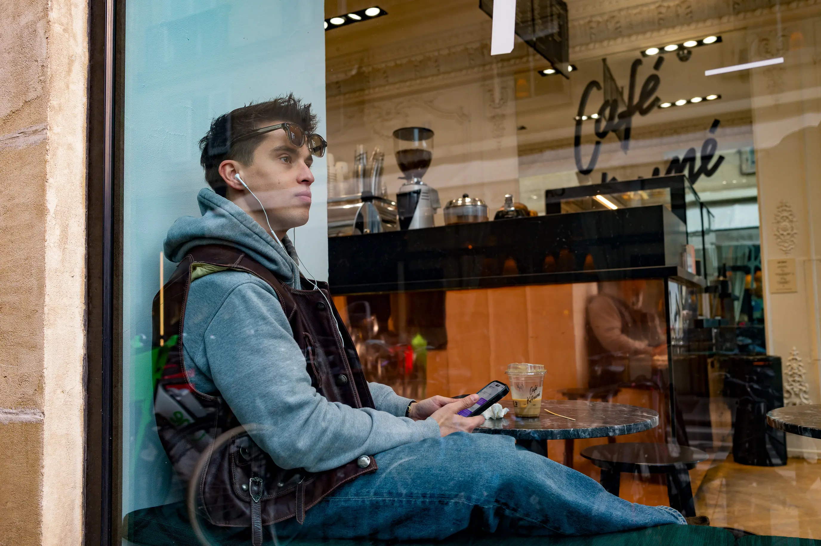 Un jeune homme assis derrière la vitre d'un café regarde au loin, son téléphone à la main