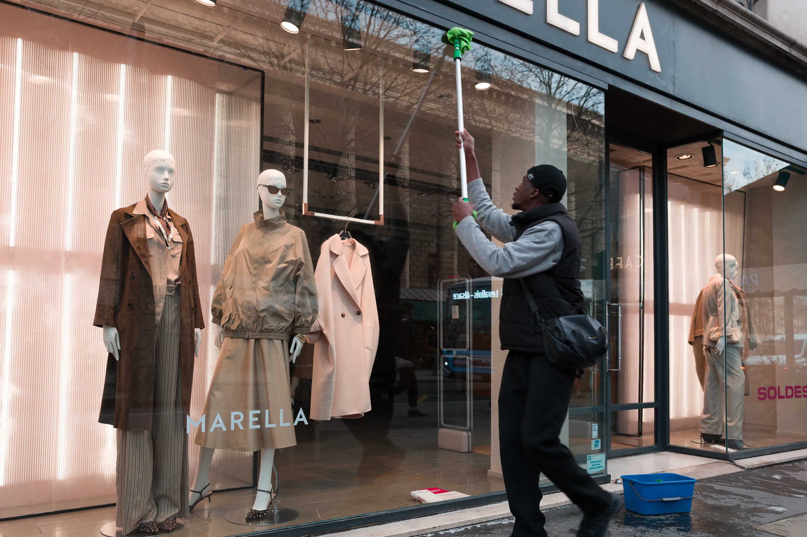 Un laveur de vitres nettoie la façade d'une boutique de mode tandis que trois mannequins élégamment vêtus semblent observer son travail derrière la vitre.
