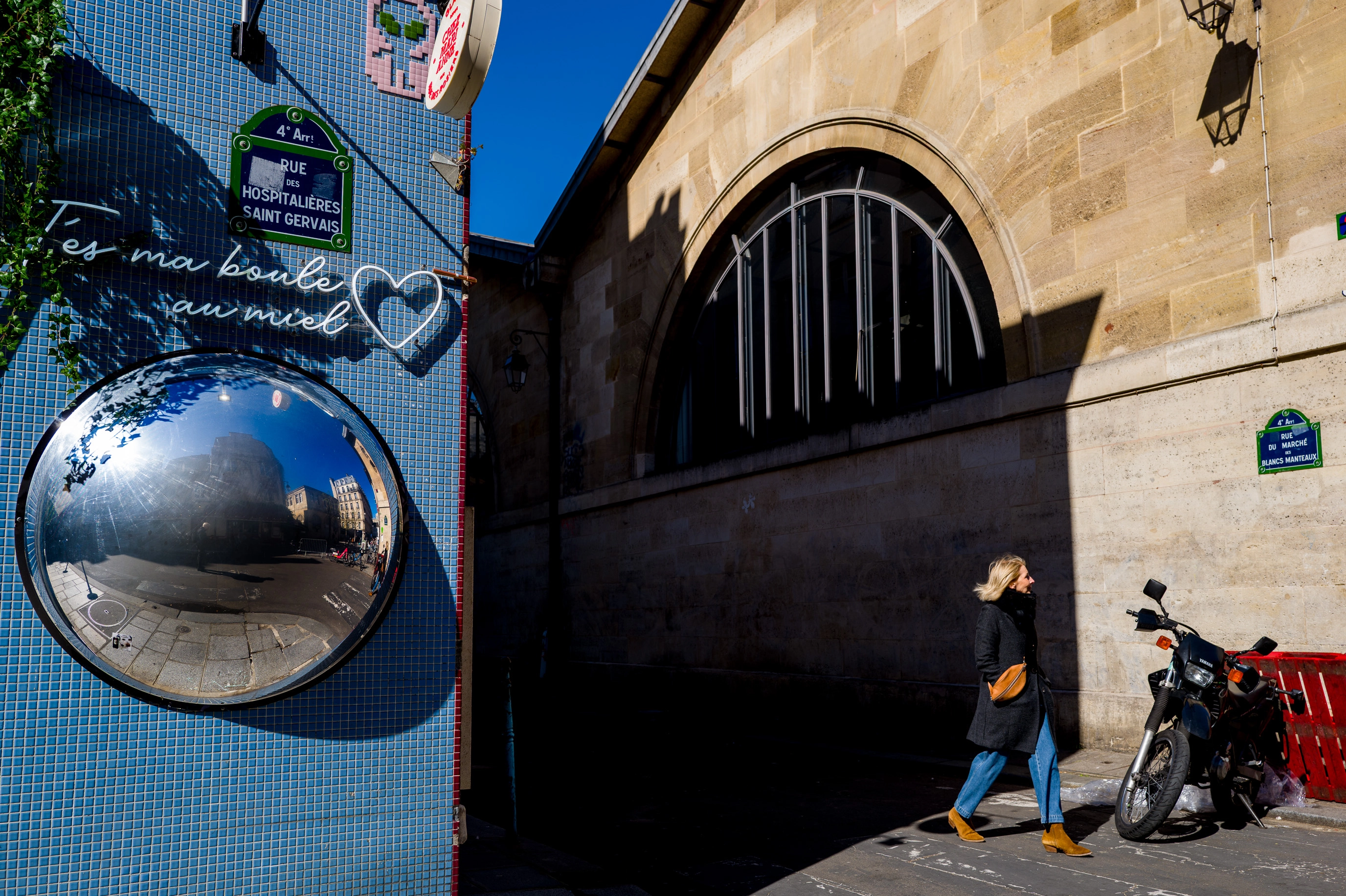 Deux façades éclairées encadrent un passage sombre ; une femme entre dans la lumière tandis qu’un miroir convexe reflète le soleil et la rue