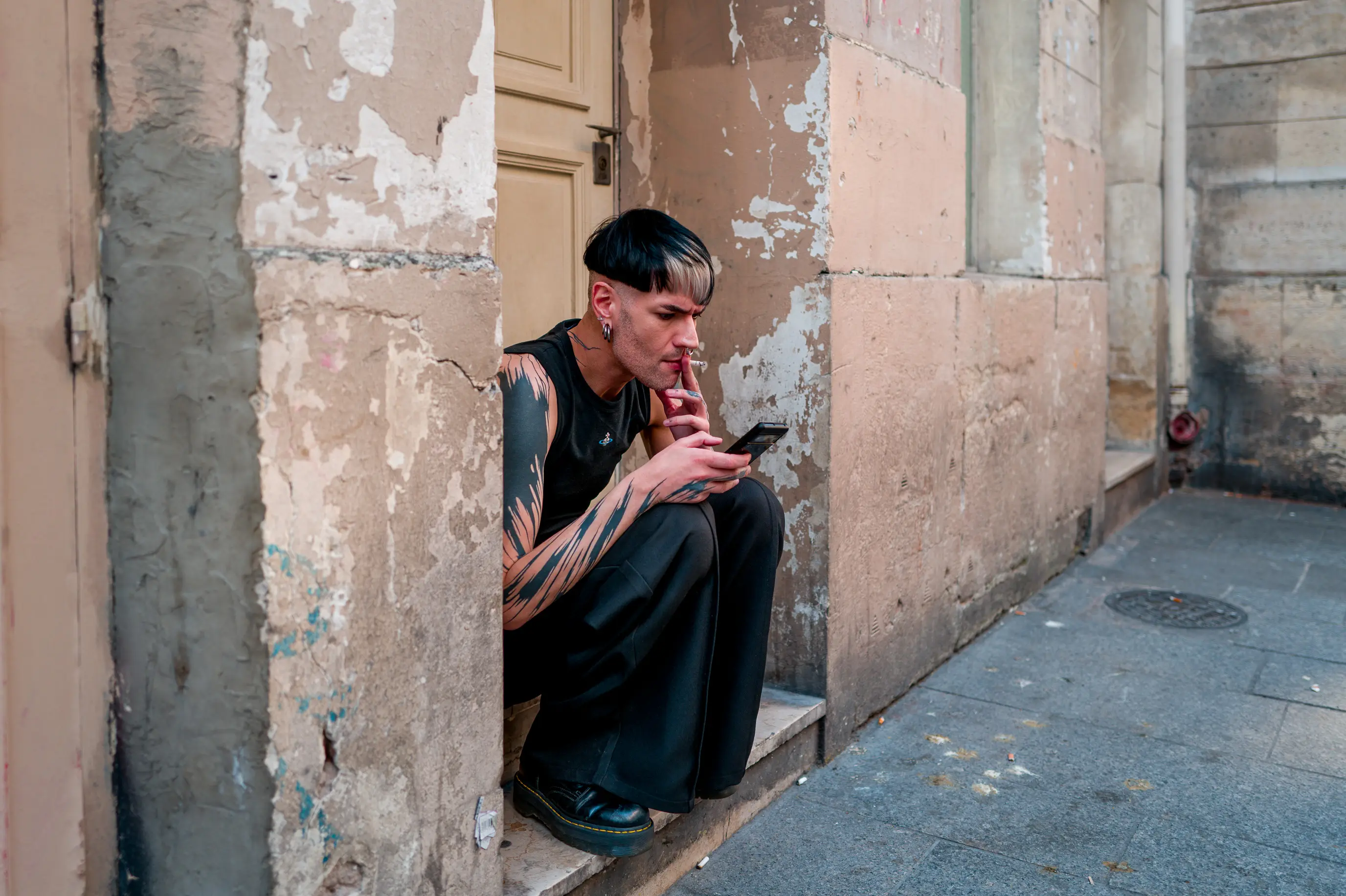 Un homme accroupi dans une embrasure semble traverser un mur, cigarette aux lèvres et téléphone à la main