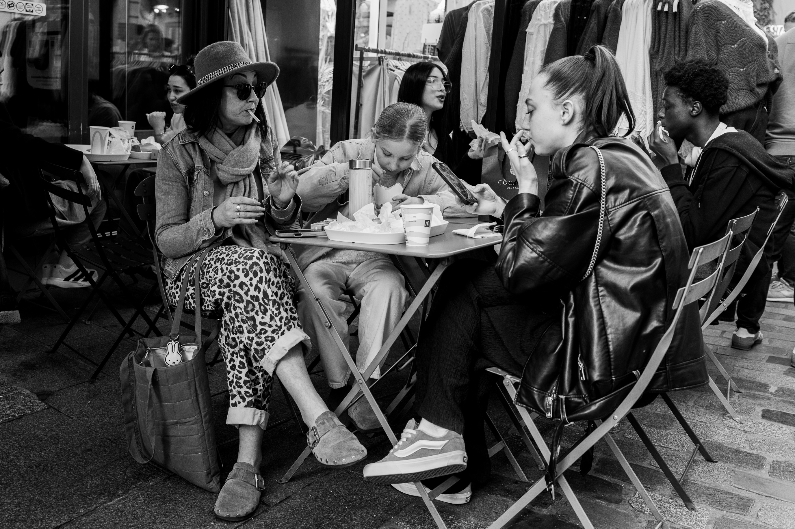 Une mère et ses deux filles partagent un repas en terrasse, entre rires et complicité, dans l’ambiance animée d’un café urbain.