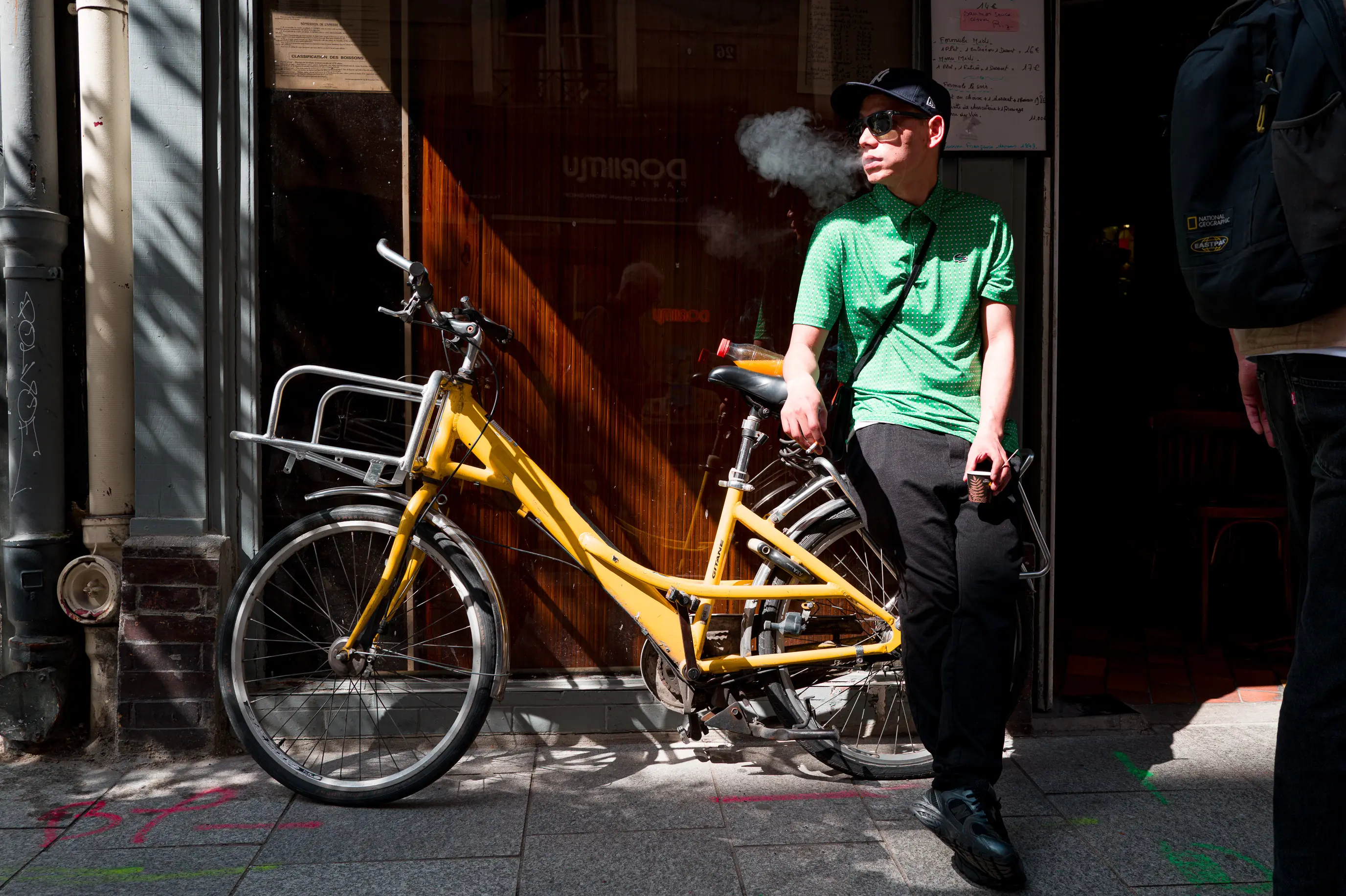 Un homme attend devant un café, appuyé contre son vélo, une cigarette à la main et le regard tourné vers la rue