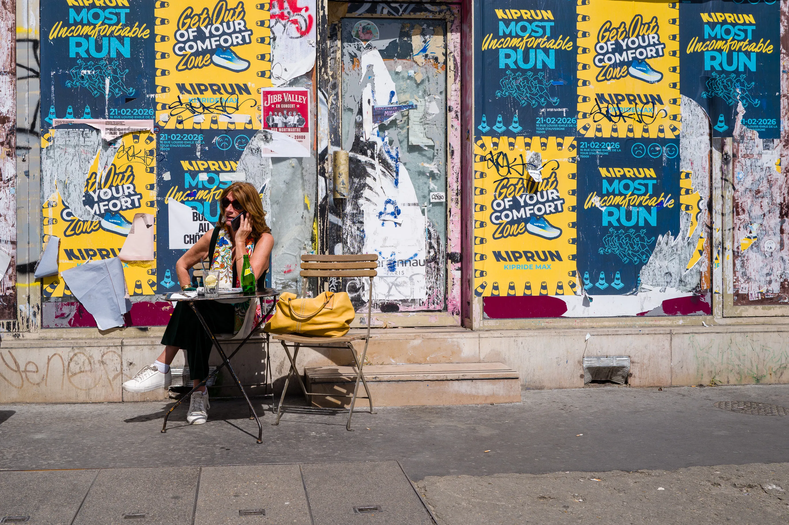 Une femme assise seule à une table légèrement écartée de la terrasse d'un café, devant un mur couvert d'affiches jaunes et bleues invitant à sortir de sa zone de confort.