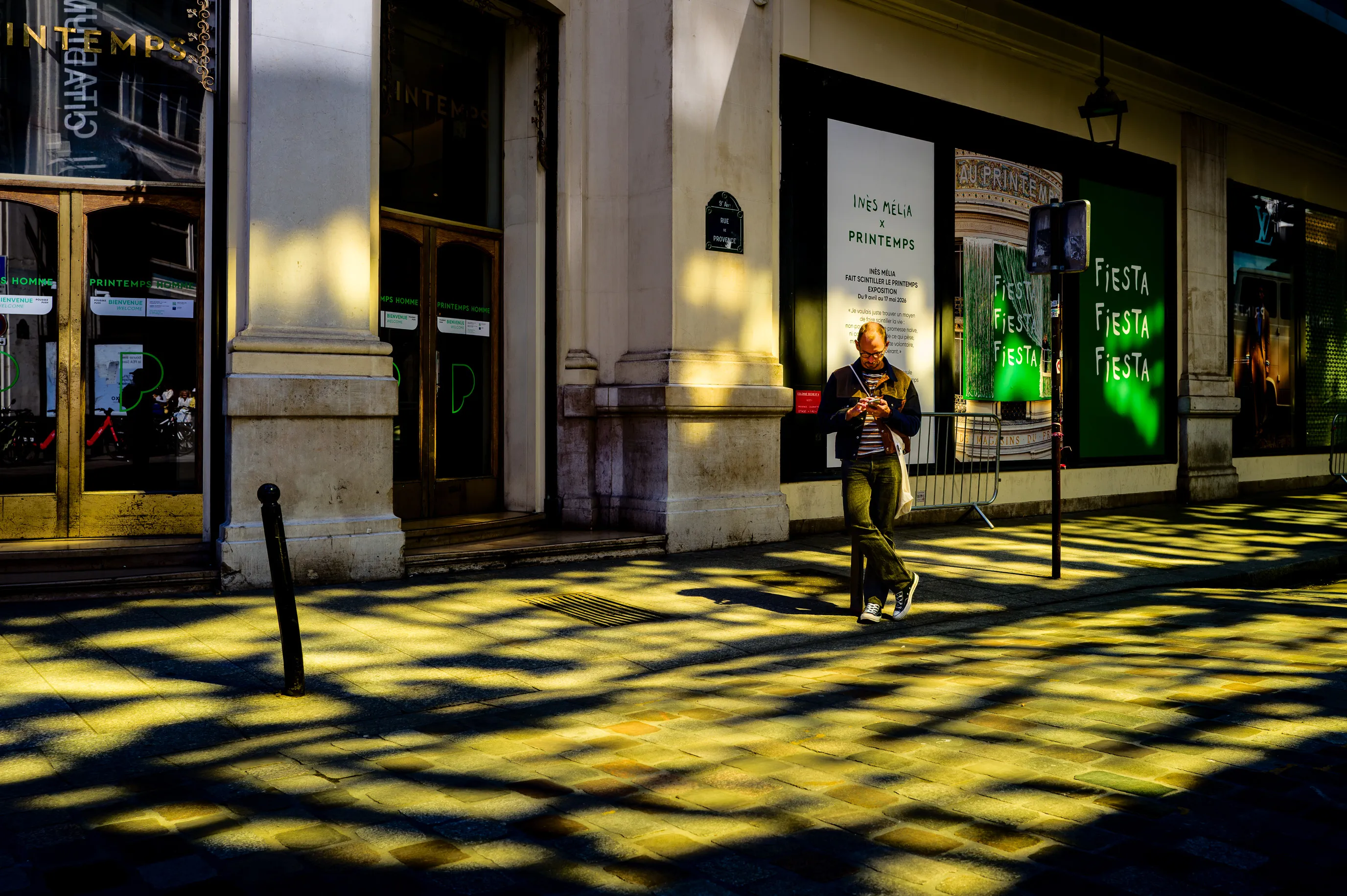 Un homme traverse des taches de lumière projetées sur un trottoir par les reflets d'une façade vitrée dorée, dans une rue de Paris.