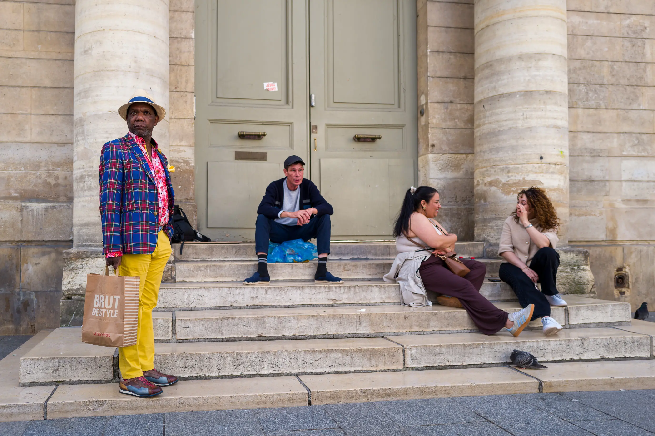 Quatre personnes occupent les marches d'un bâtiment, chacune dans une attitude différente, devant une grande porte encadrée de colonnes