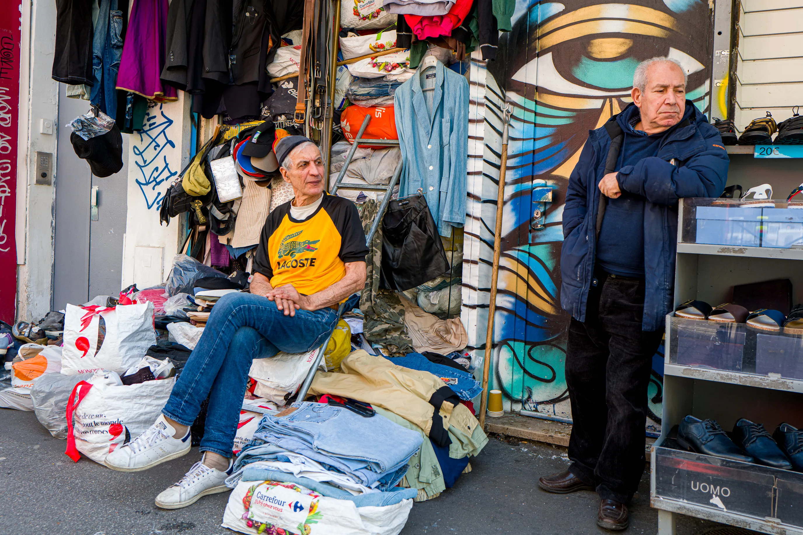 Deux hommes devant une boutique de vêtements d'occasion rue du Faubourg du Temple à Paris. L'un est assis au milieu de sa marchandise étalée sur le trottoir tandis que l'autre est adossé à un présentoir, sous une grande fresque représentant un œil.