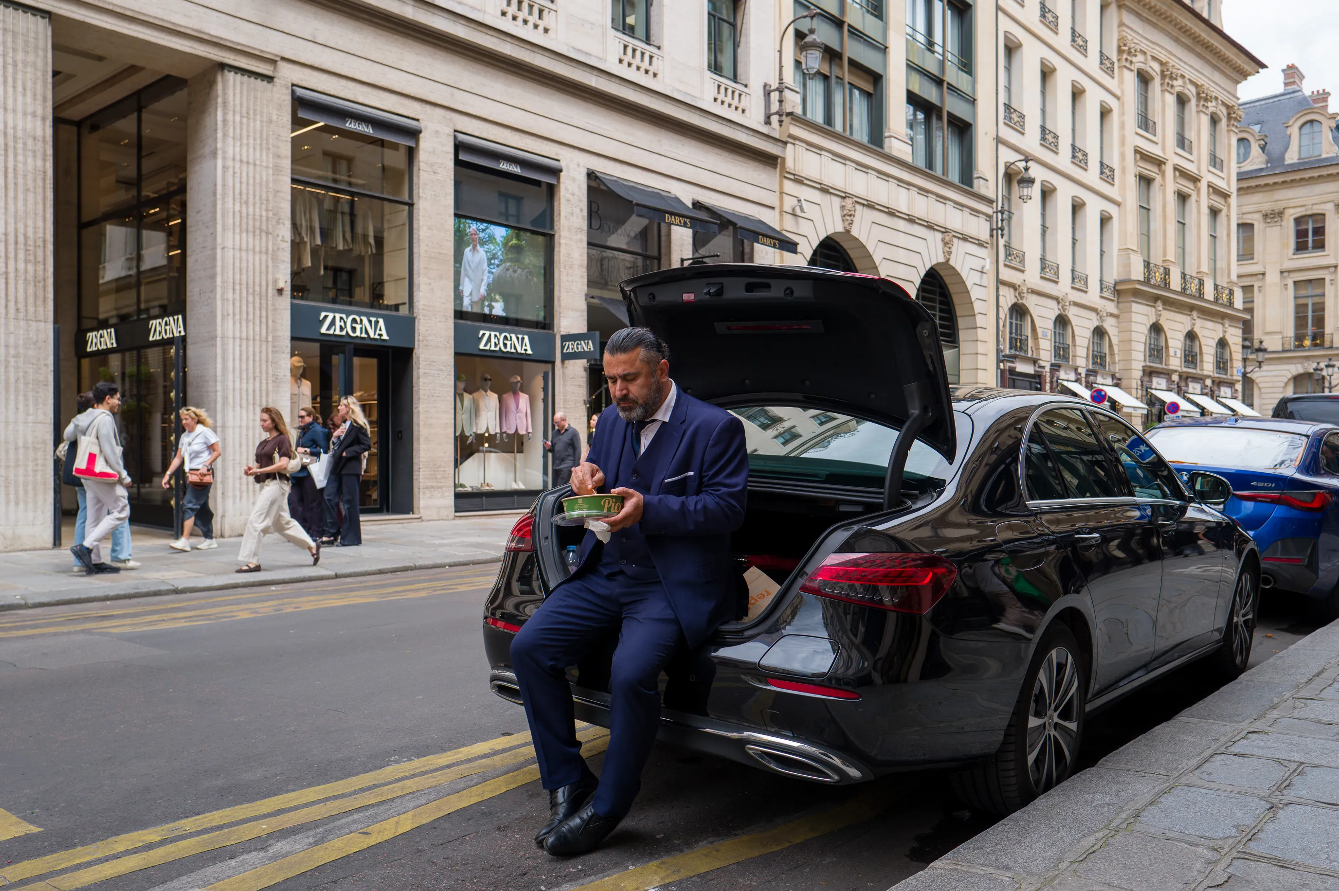 Un homme en costume bleu marine, assis sur le bord du coffre ouvert d’une Mercedes noire, mange dans un plat préparé dans une boîte en plastique. Rue Saint-Honoré, devant Zegna, les passants passent, les vitrines brillent. Lui reste immobile, le bol à la main, indifférent au luxe qui l’entoure. La voiture attend, le moteur éteint.