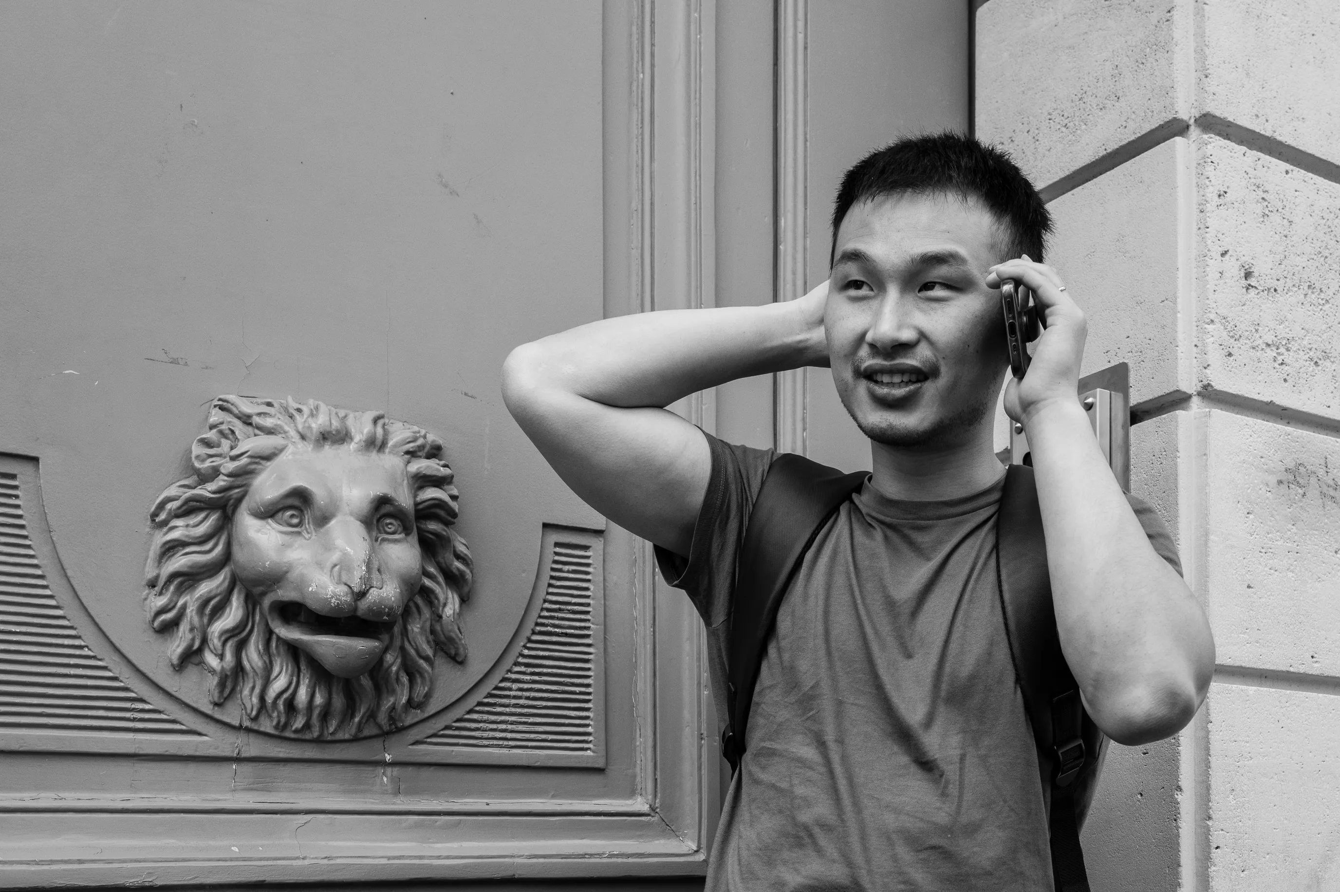 Un homme au téléphone devant une porte ornée d'une tête de lion en bas-relief, en noir et blanc.