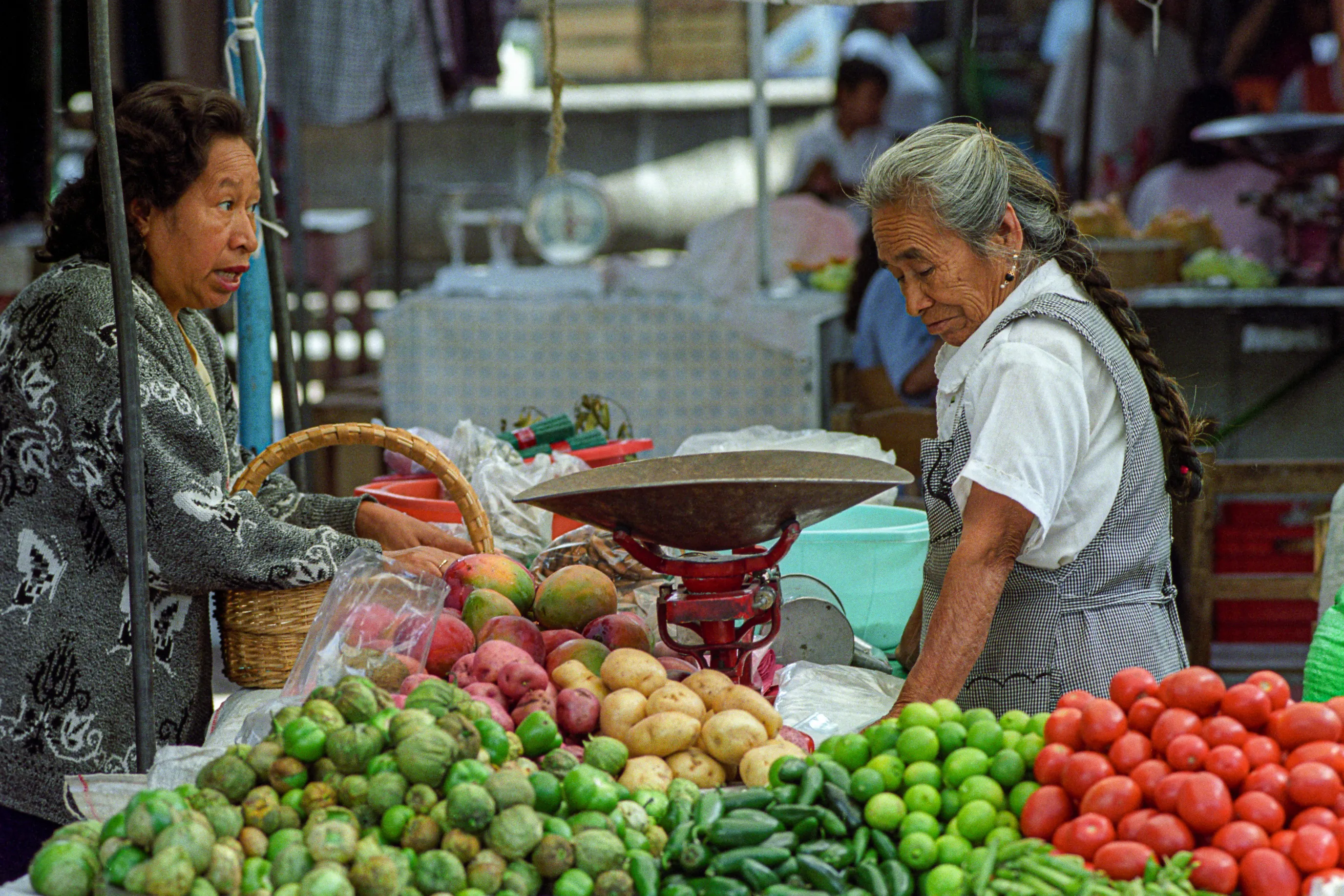 Au marché de Tepoztlán, une cliente négocie le prix de tomates avec une marchande derrière un étal coloré de fruits et de légumes.