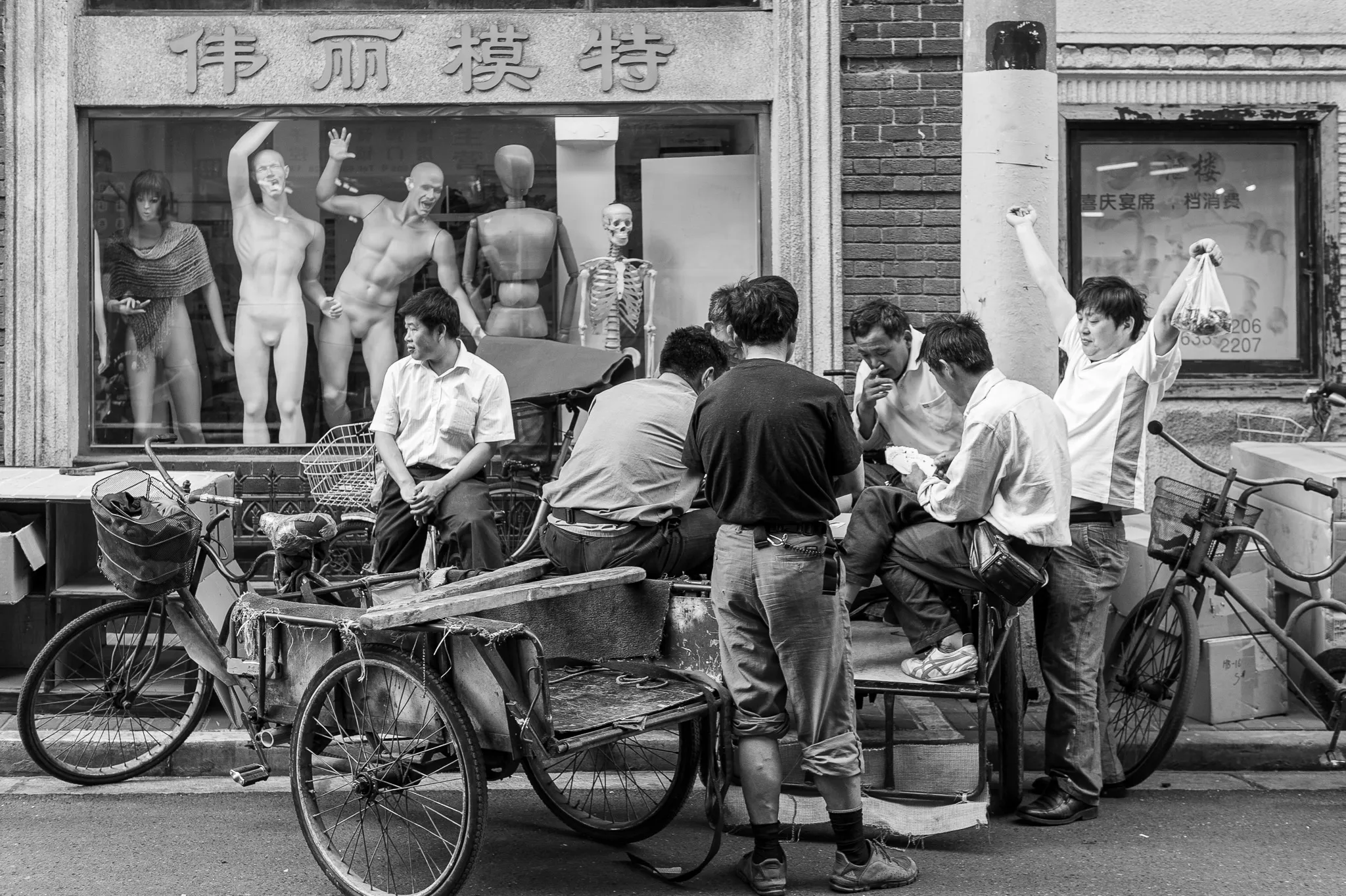 Scène de rue à Shanghai en 2011 : des ouvriers se reposent et discutent devant une vitrine remplie de mannequins figés et d’un squelette.