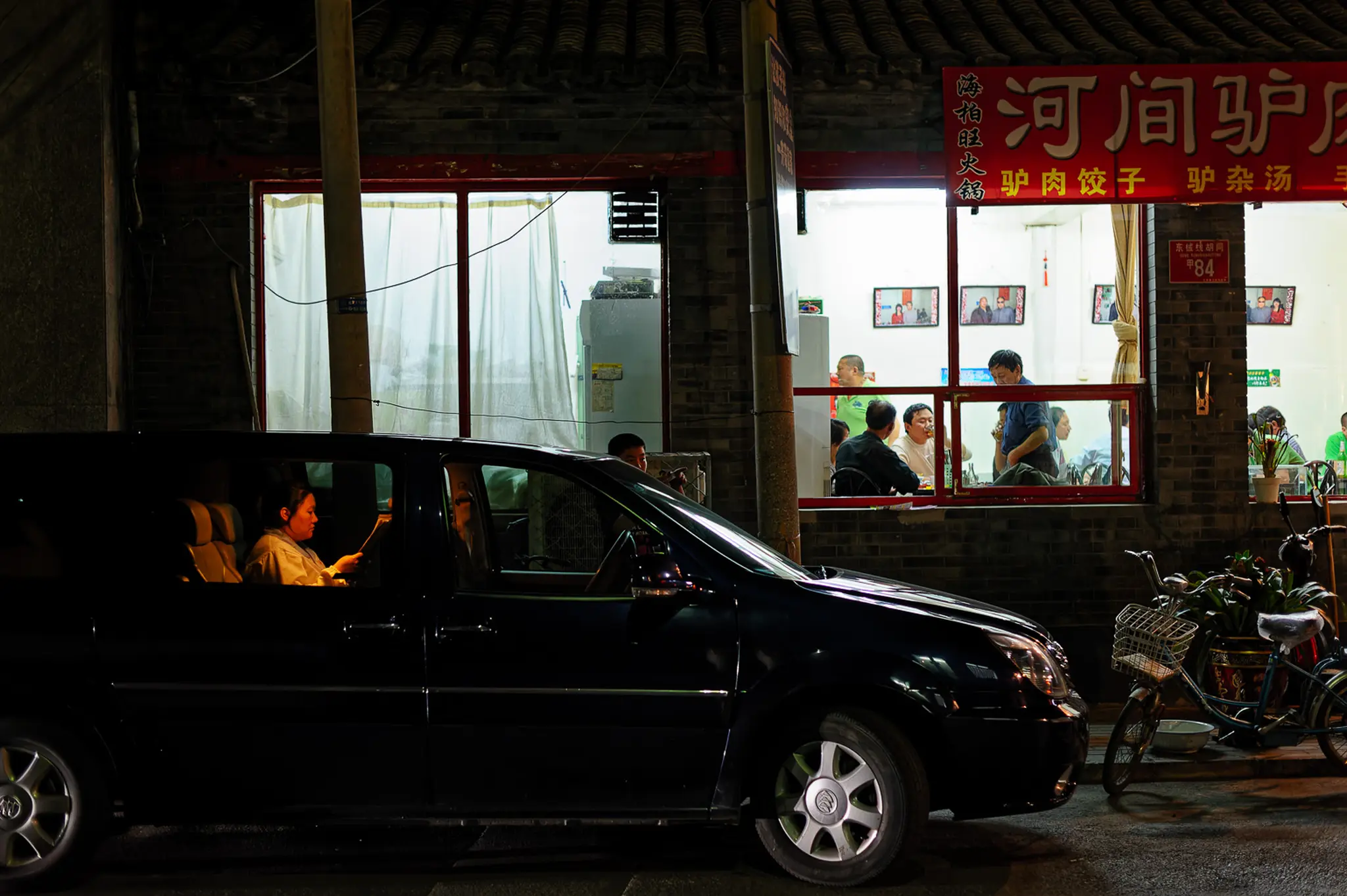De nuit, une voiture noire est arrêtée devant un restaurant très éclairé ; dans l’ombre de l’habitacle, une passagère regarde son téléphone