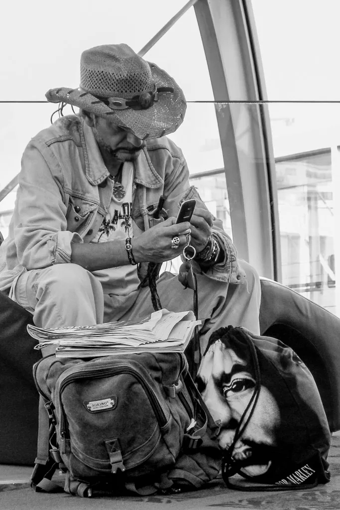 Homme assis, concentré sur son téléphone, un sac imprimé d’un grand visage posé à ses pieds