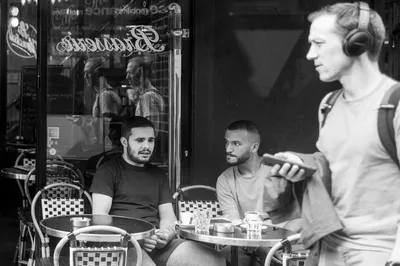 Deux hommes assis à une terrasse tandis qu’un passant traverse la scène, téléphone en main