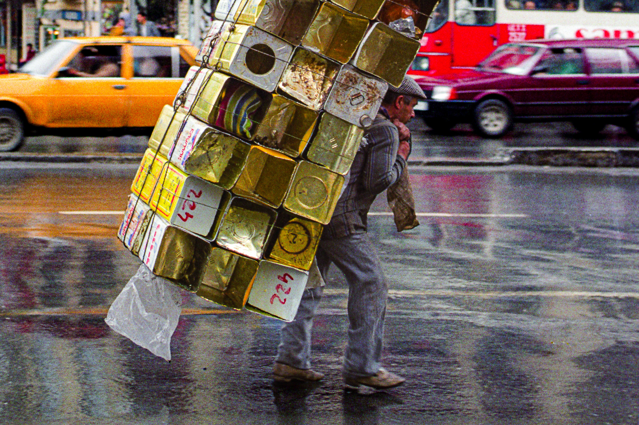 Un porteur ploie sous une pile de bidons d’huile vides, avançant dans une rue mouillée devant un bus rempli de passagers