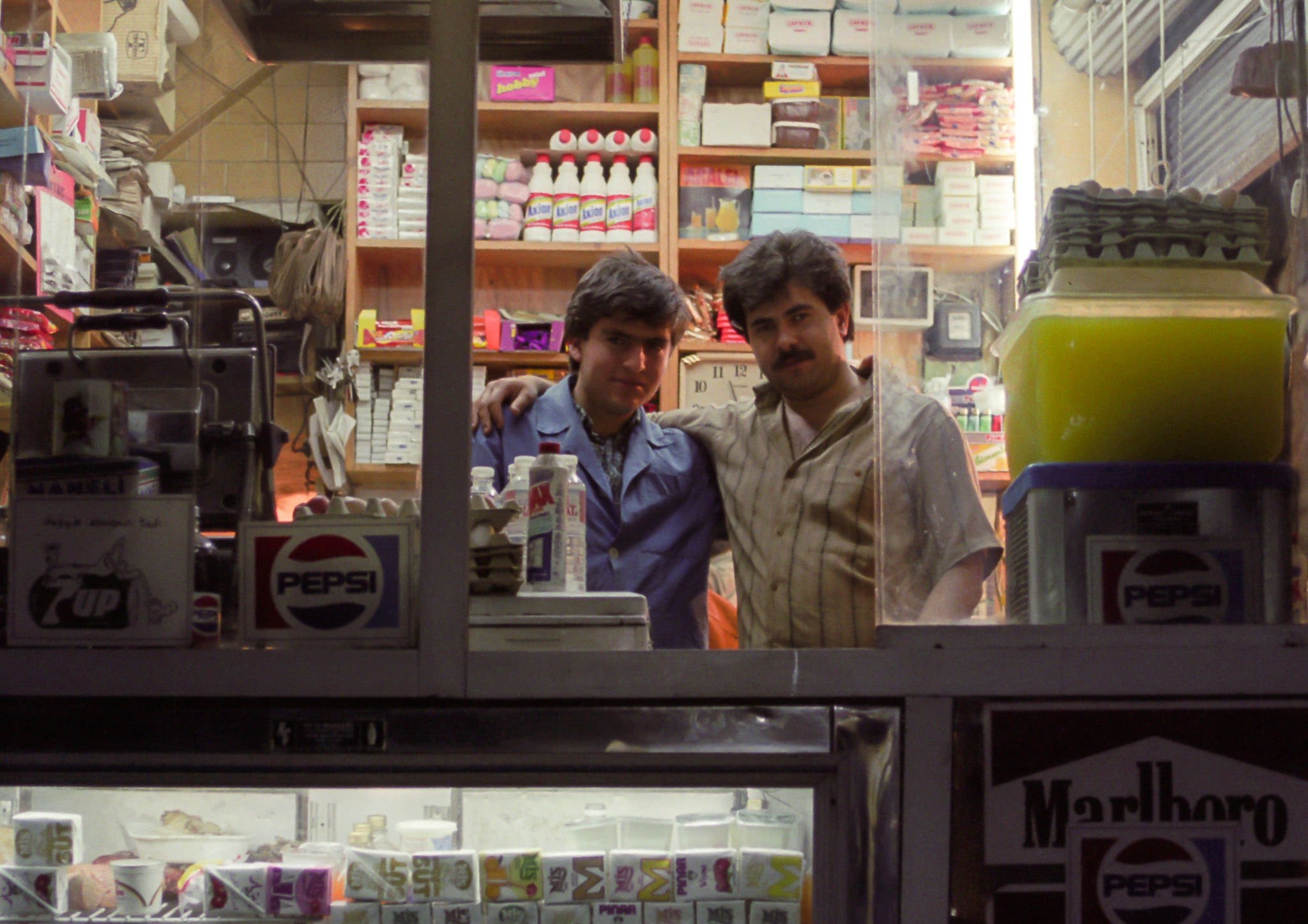 Deux jeunes épiciers posent derrière la vitrine de leur boutique éclairée, entourés de produits de consommation courante à Istanbul en 1990.