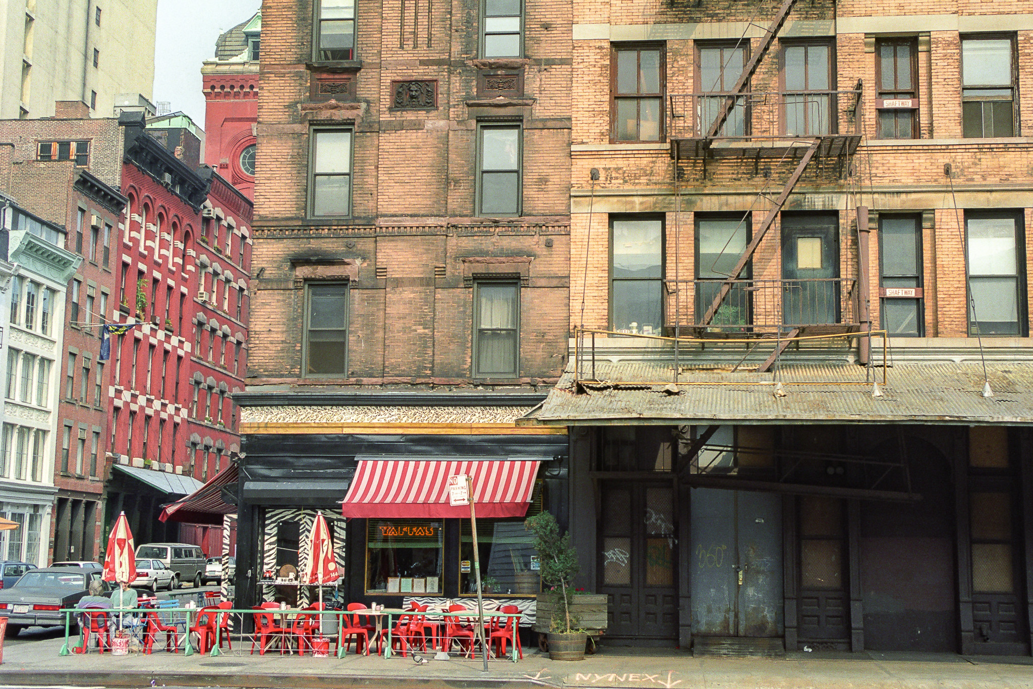 Rue de Hell’s Kitchen à New York en 1995, façades de briques anciennes, restaurant avec terrasse de chaises rouges.