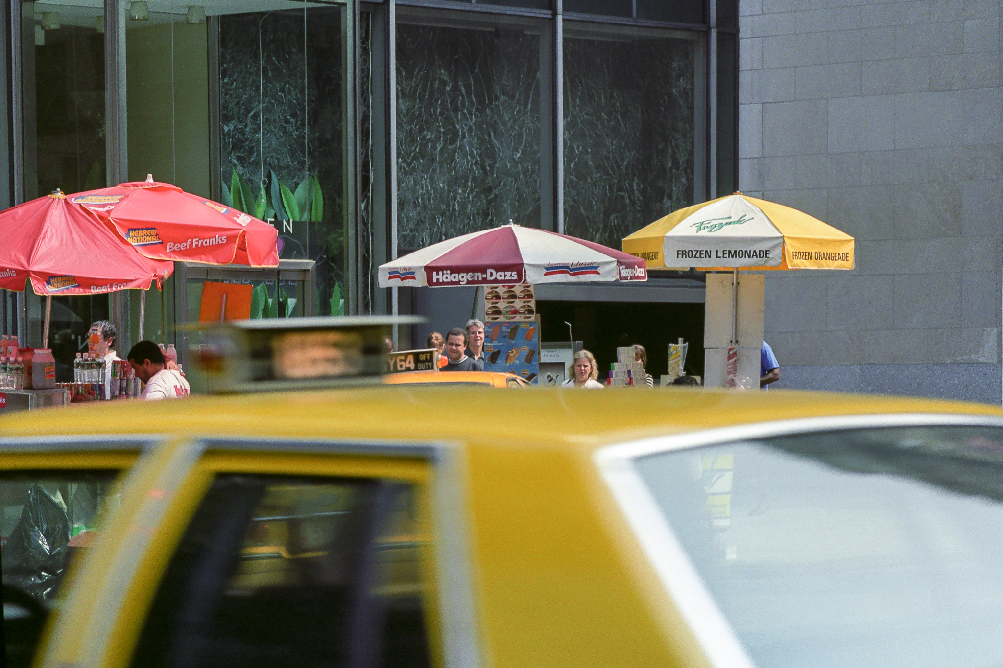 Scène de rue à New York en 1995, parasols de stands de hot-dogs et glaces, taxi jaune passant au premier plan.