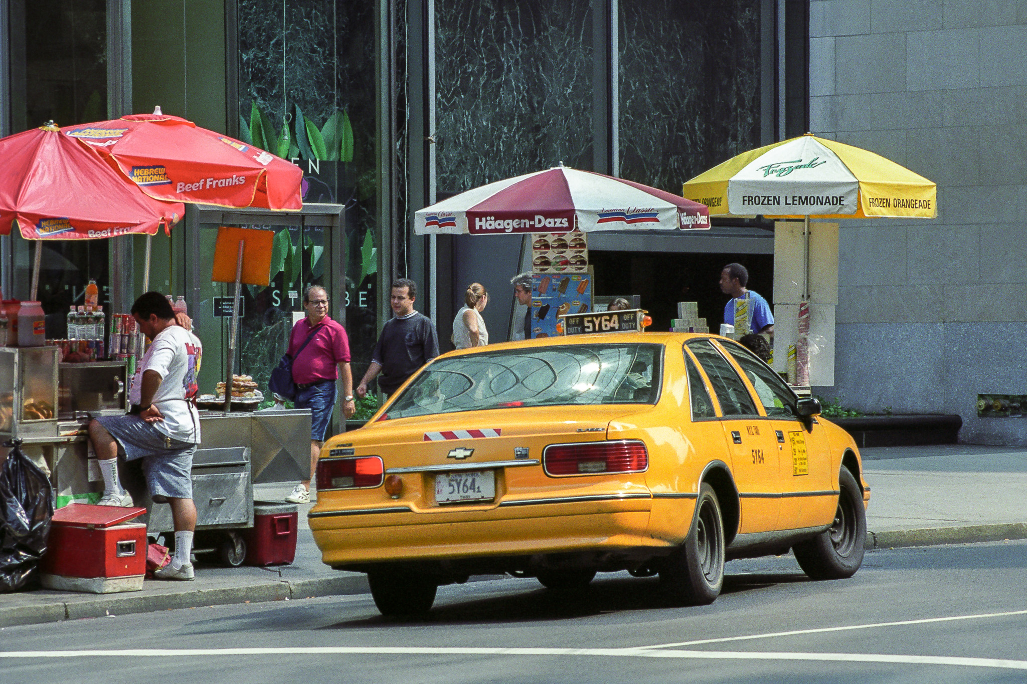 Scène de rue à New York en 1995, parasols de stands de hot-dogs et glaces, taxi jaune passant au premier plan.
