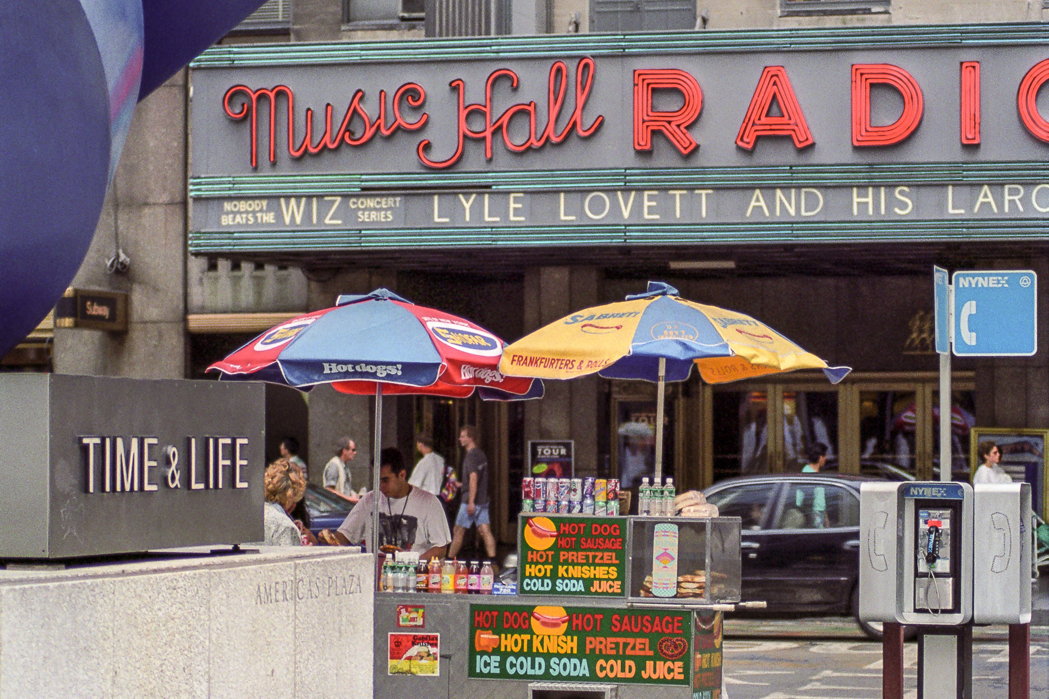 Stand de hot-dogs devant le Radio City Music Hall à New York en 1995, avec parasols colorés et téléphones publics.