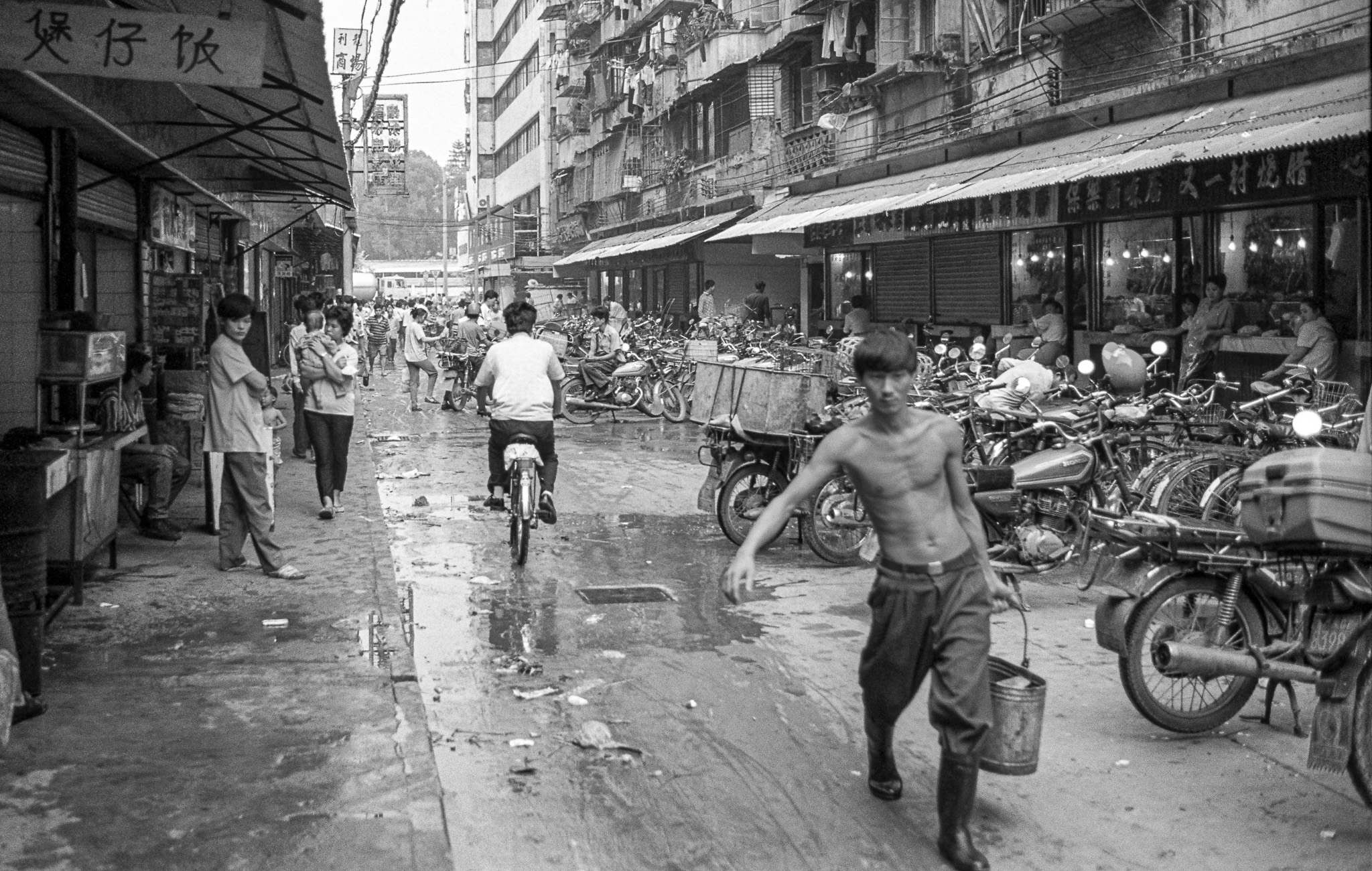 Scène de rue à Canton en 1995, jeune homme torse nu avec un seau, rue humide bordée de motos et de vélos, habitants en mouvement.