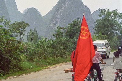 Un cycliste portant un grand drapeau rouge sur une route bordée de pitons karstiques.