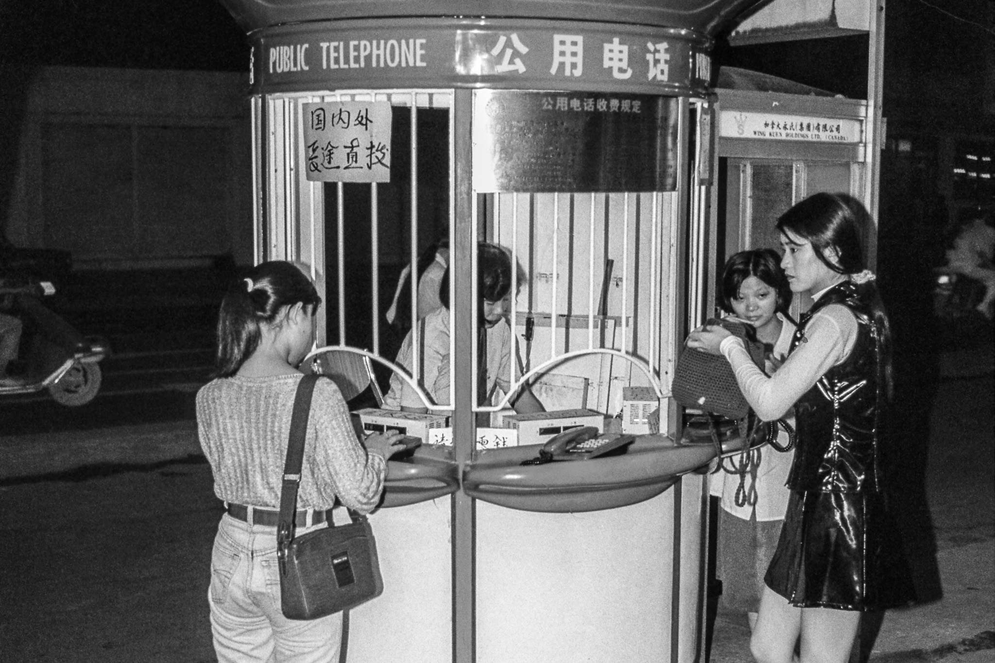 Scène nocturne à Canton en 1995 : des femmes utilisent une cabine téléphonique publique gérée par un guichetier, le téléphone personnel n’étant pas encore courant.