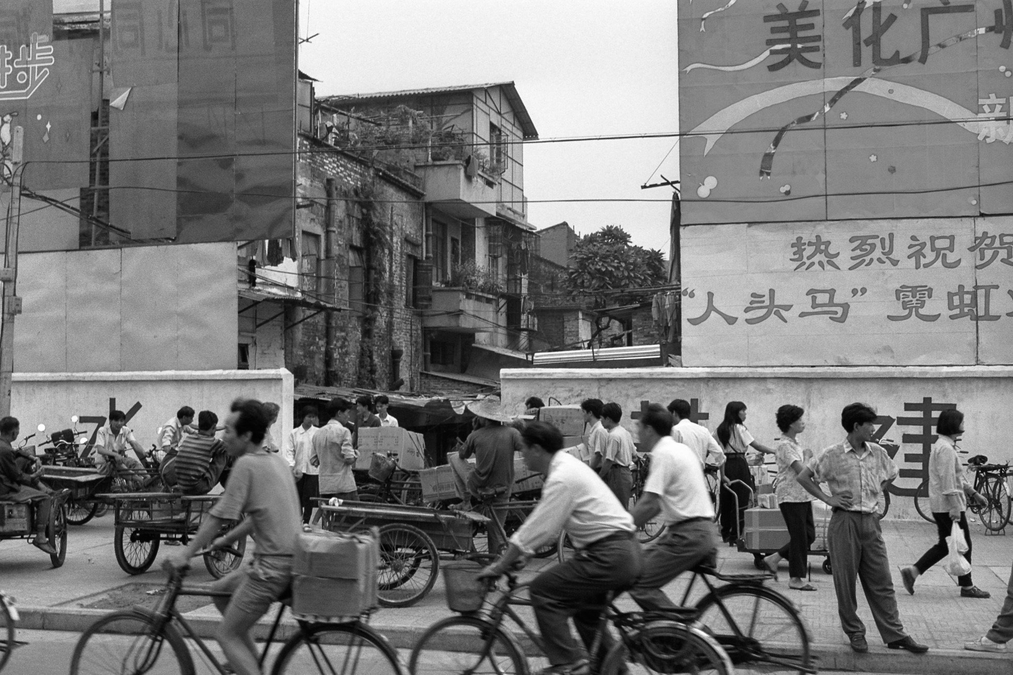 Scène de rue à Canton en 1995, vélos et triporteurs chargés de caisses circulant parmi les passants, avec en arrière-plan des façades anciennes et une grande affiche murale.