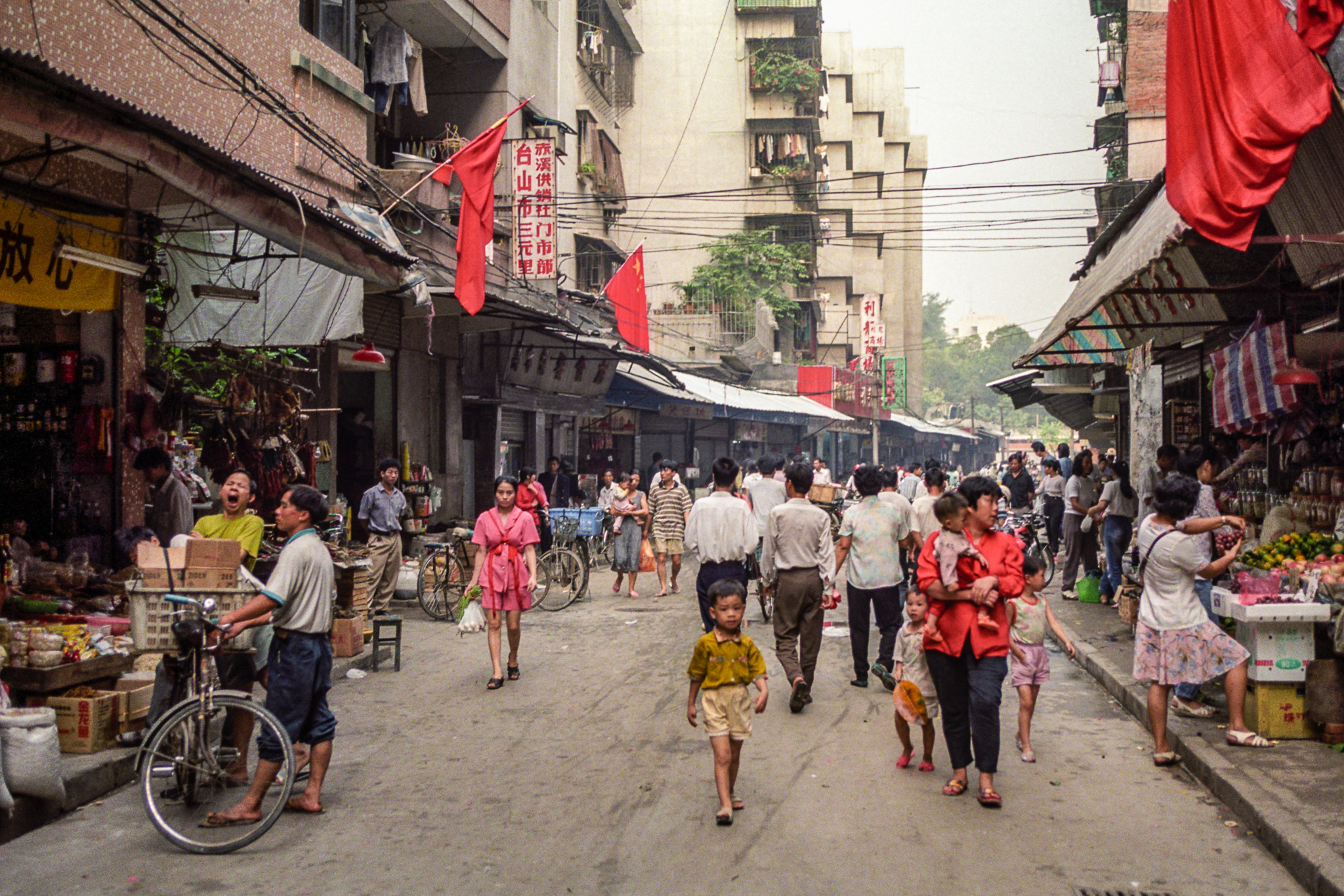 Scène de rue à Canton en 1995, marché populaire avec passants, enfants, vélos et échoppes animées des deux côtés.