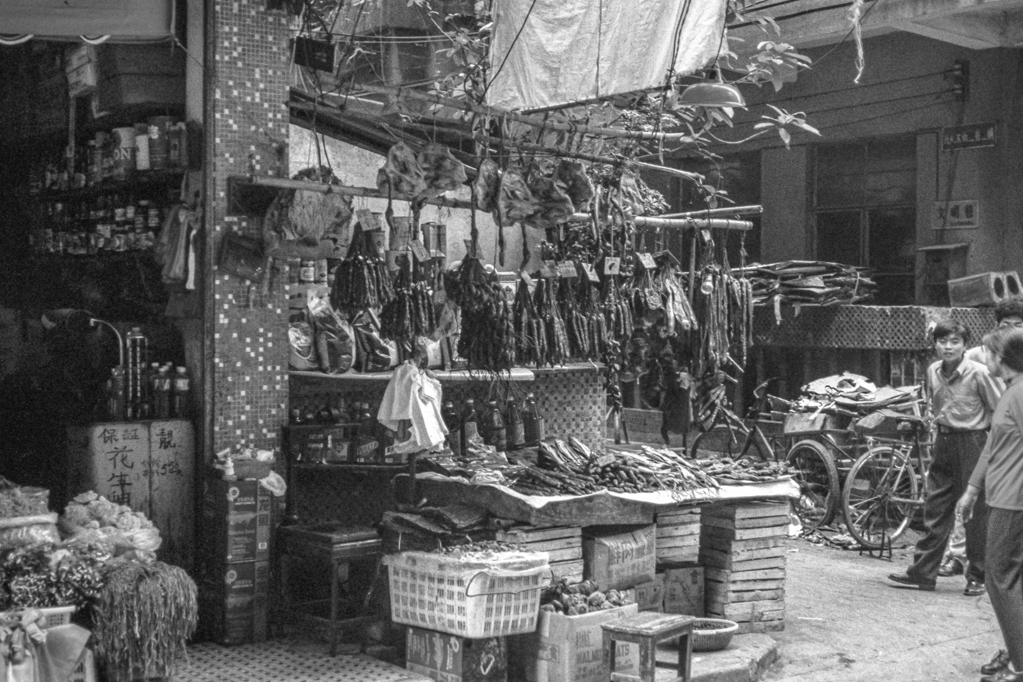 Étal de marché à Canton en 1995, mélange d’ordre et de désordre avec légumes, saucisses suspendues, paniers et produits variés.