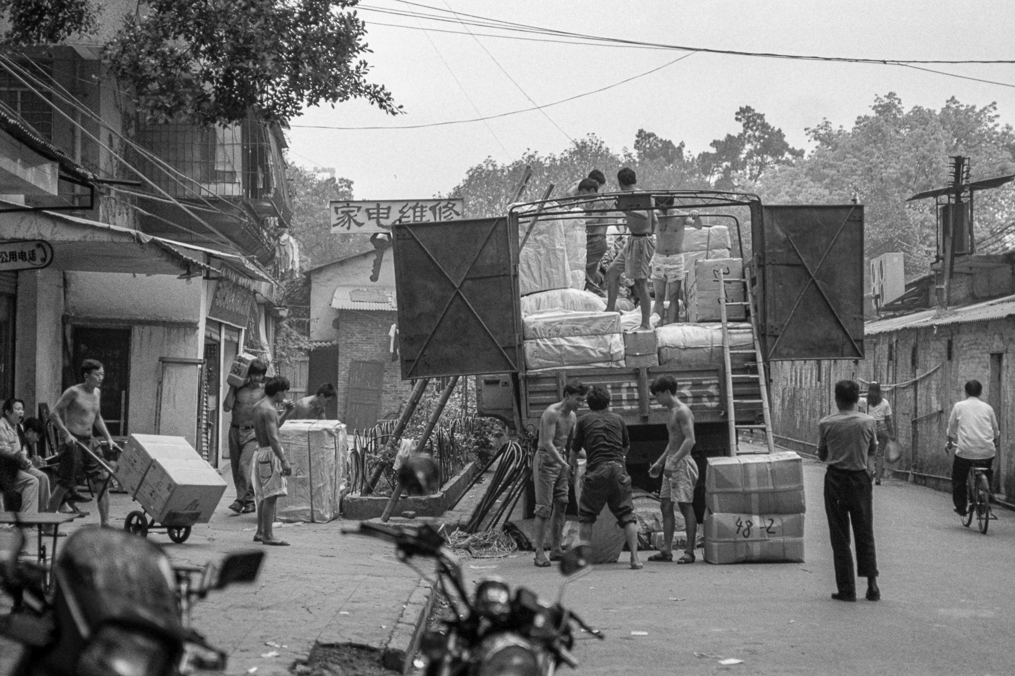 Scène de rue à Canton en 1995 : plusieurs hommes torse nu déchargent un camion rempli de marchandises, cartons et ballots s’accumulent sur la chaussée.