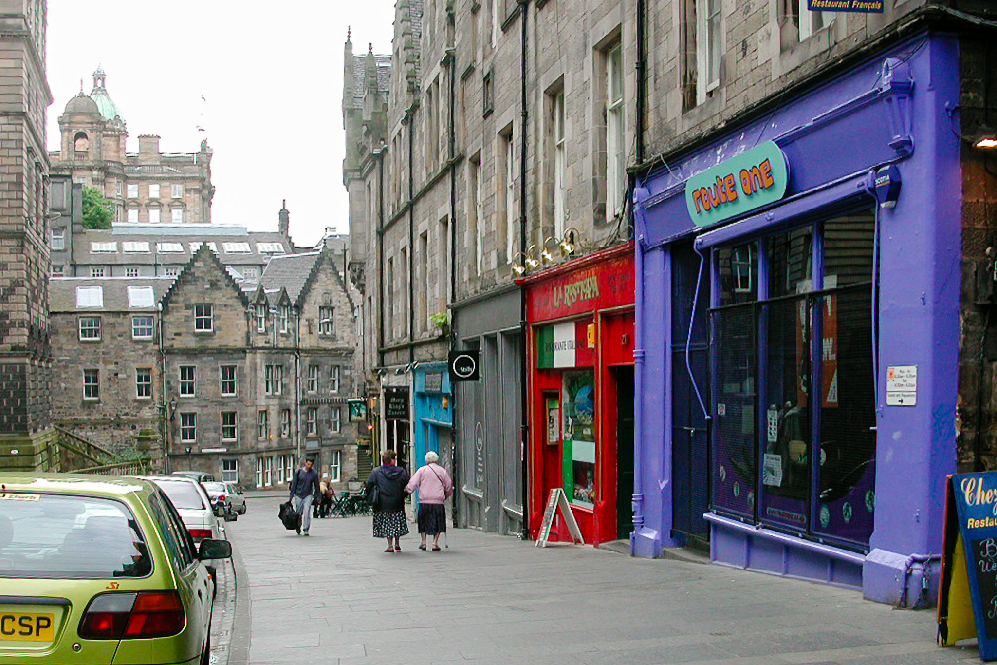 Édimbourg, rue en pente de la vieille ville : façades en pierre grise, boutiques violette et rouge, deux passantes de dos, voitures garées à gauche.