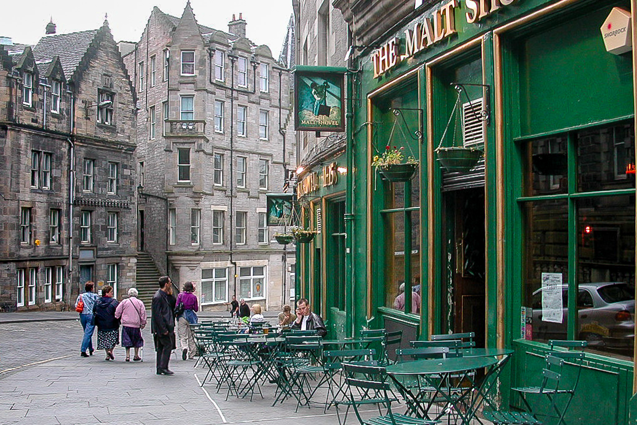 Édimbourg, pub The Malt Shovel : façade verte, tables en terrasse vides ou occupées, passants dans une rue bordée d’immeubles de pierre grise.