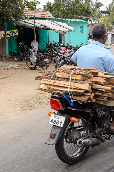 Une moto transporte une lourde pile de planches dans une rue de village.