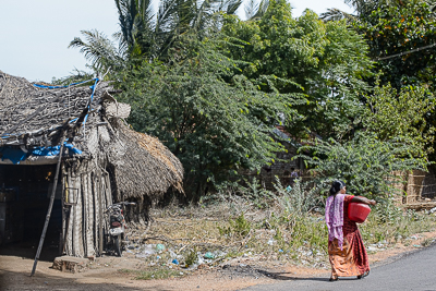 Une femme marche devant une hutte de palmes adossée à la végétation.