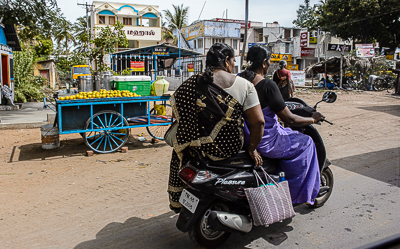 Deux femmes sur un scooter dépassent une charrette bleue chargée de marchandises.