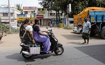 Le scooter et ses deux passagères poursuivent leur chemin au milieu des camions et des commerces.